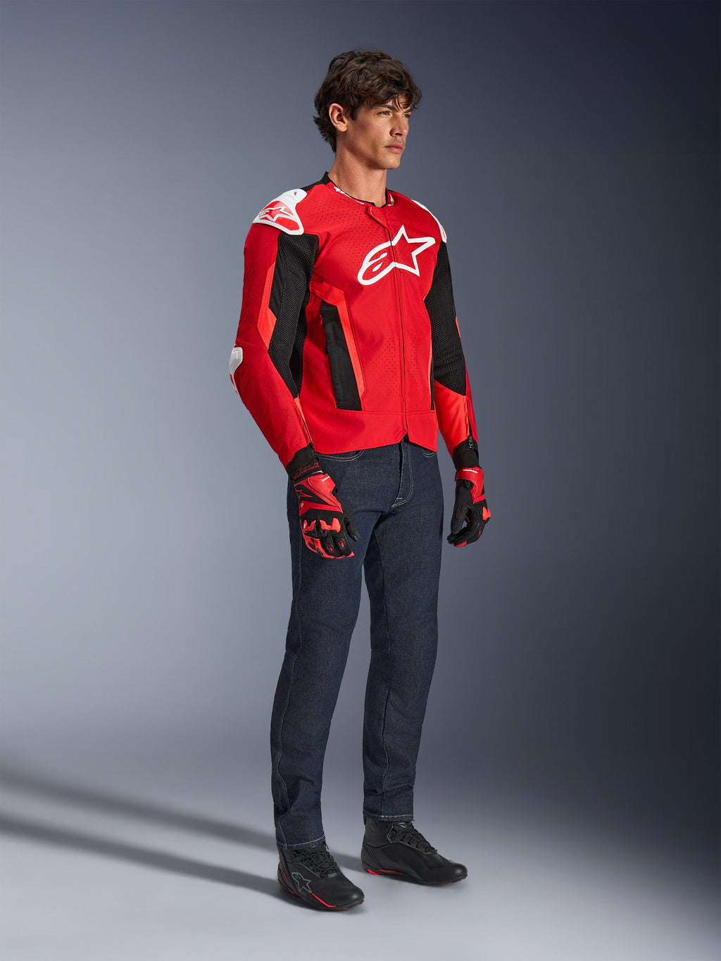 Chaqueta Alpinestars T-GP Air, chaqueta deportiva para moto, rojo brillante y negro con detalles en blanco, usada por un modelo con jeans azul oscuro y guantes de carreras rojos, con paneles de malla de alto flujo y deslizadores de hombro blancos, ajuste deportivo aerodinámico para conducir en climas cálidos