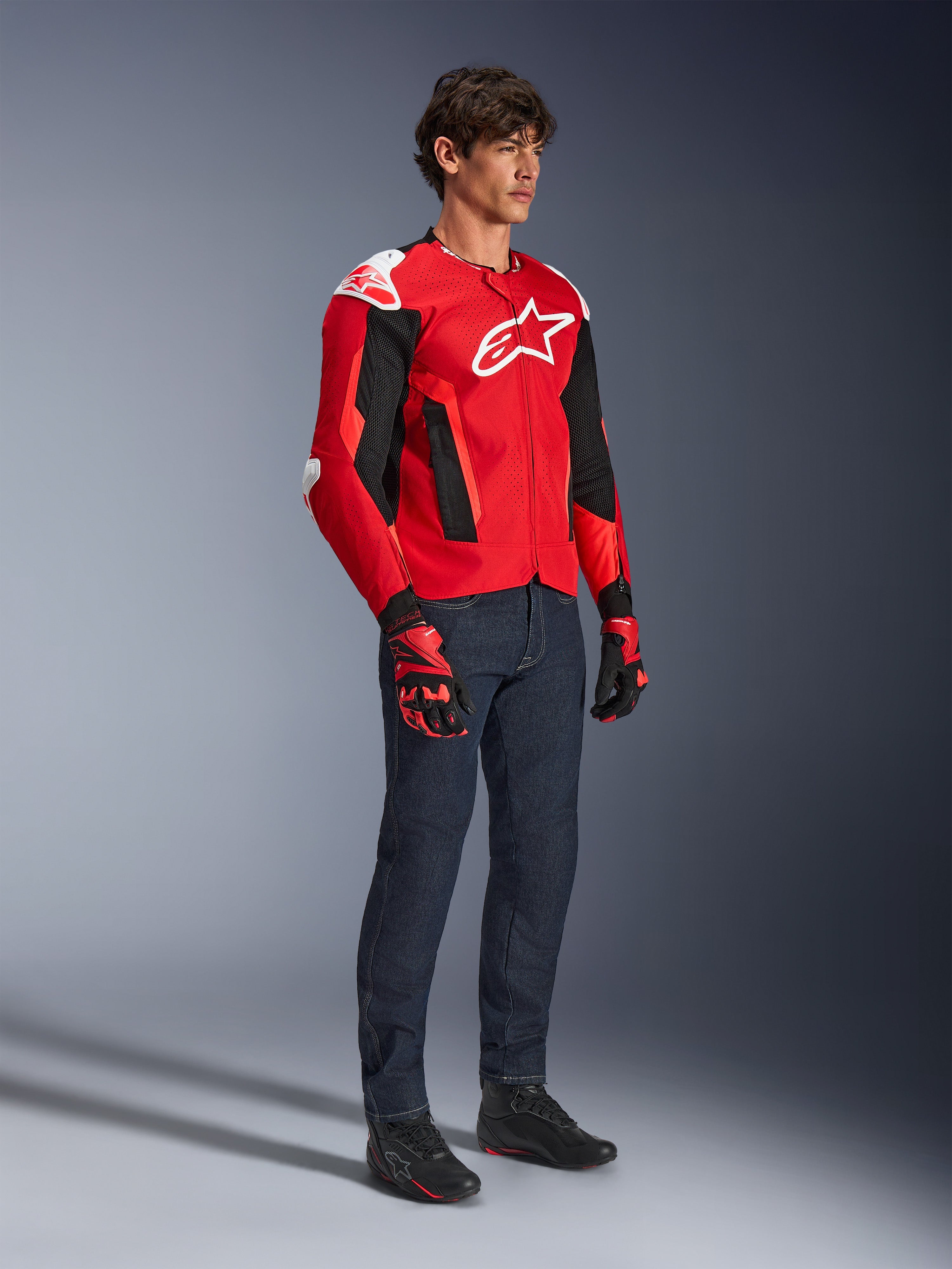Chaqueta Alpinestars T-GP Air, chaqueta deportiva para moto, rojo brillante y negro con detalles en blanco, usada por un modelo con jeans azul oscuro y guantes de carreras rojos, con paneles de malla de alto flujo y deslizadores de hombro blancos, ajuste deportivo aerodinámico para conducir en climas cálidos