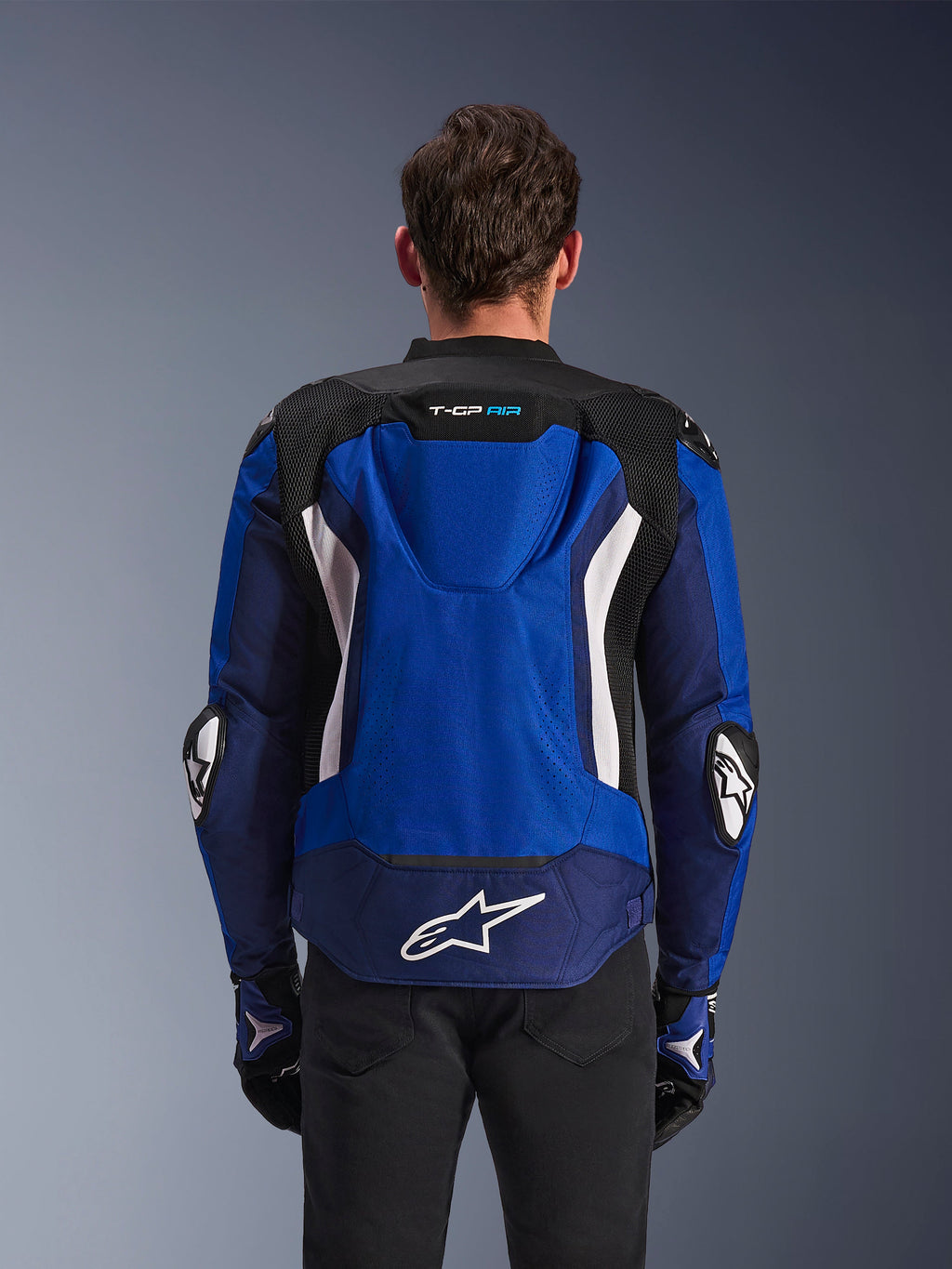Alpinestars T-GP Air Jacket, Sport Textile Motorcycle Jacket, Bright Blue Navy and White, rear view featuring the integrated aerodynamic hump and large white logo on lower back, worn with matching blue and black riding gloves