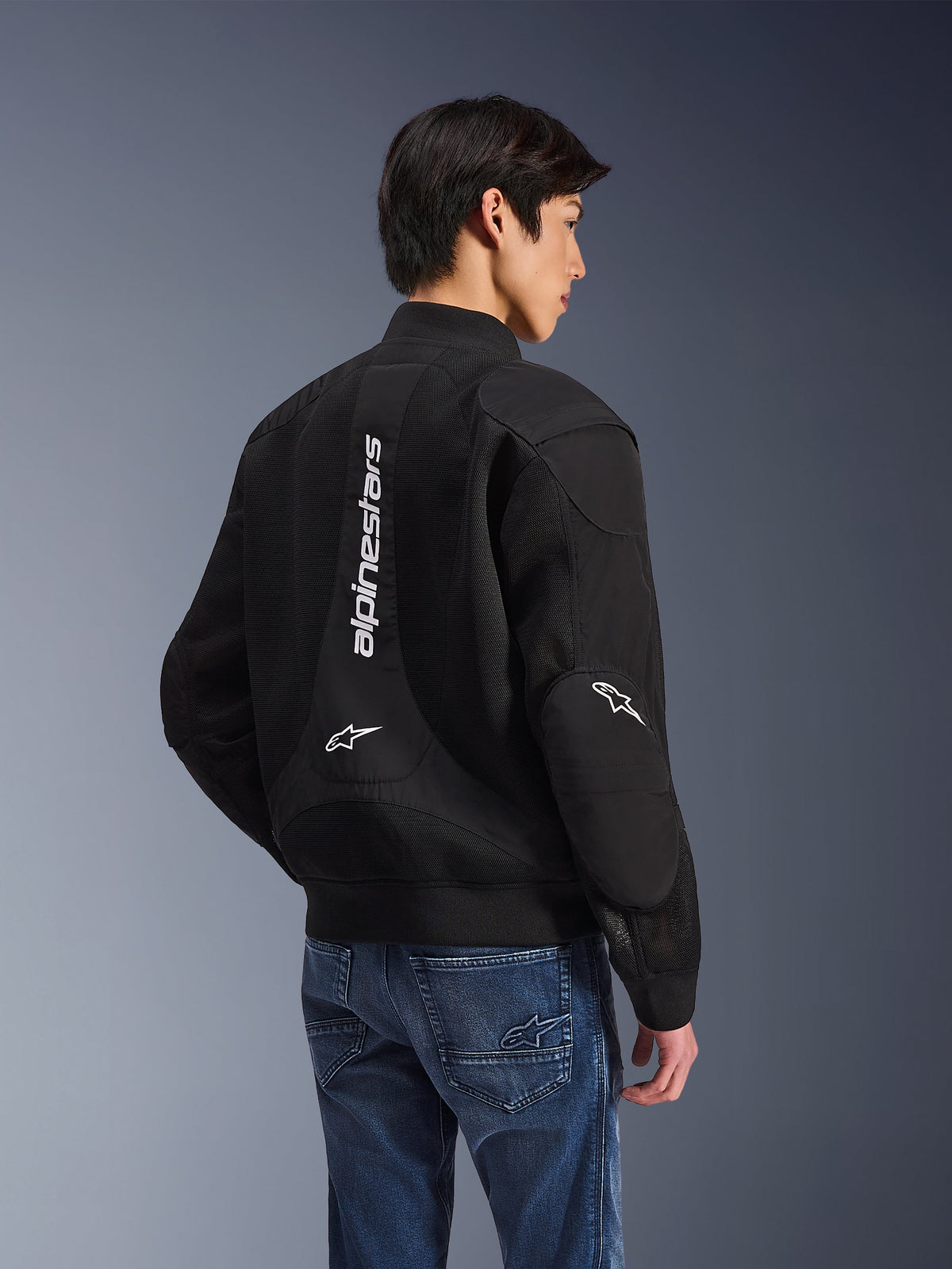 Chaqueta Alpinestars Flight Air, chaqueta de moto negra, vista posterior con un gran logotipo blanco vertical y paneles de ventilación de malla, estilo bomber con dobladillo acanalado, combinada con vaqueros azules, equipamiento de conducción urbano contemporáneo