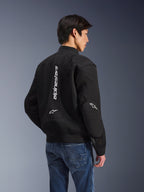Chaqueta Alpinestars Flight Air, chaqueta de moto negra, vista posterior con un gran logotipo blanco vertical y paneles de ventilación de malla, estilo bomber con dobladillo acanalado, combinada con vaqueros azules, equipamiento de conducción urbano contemporáneo