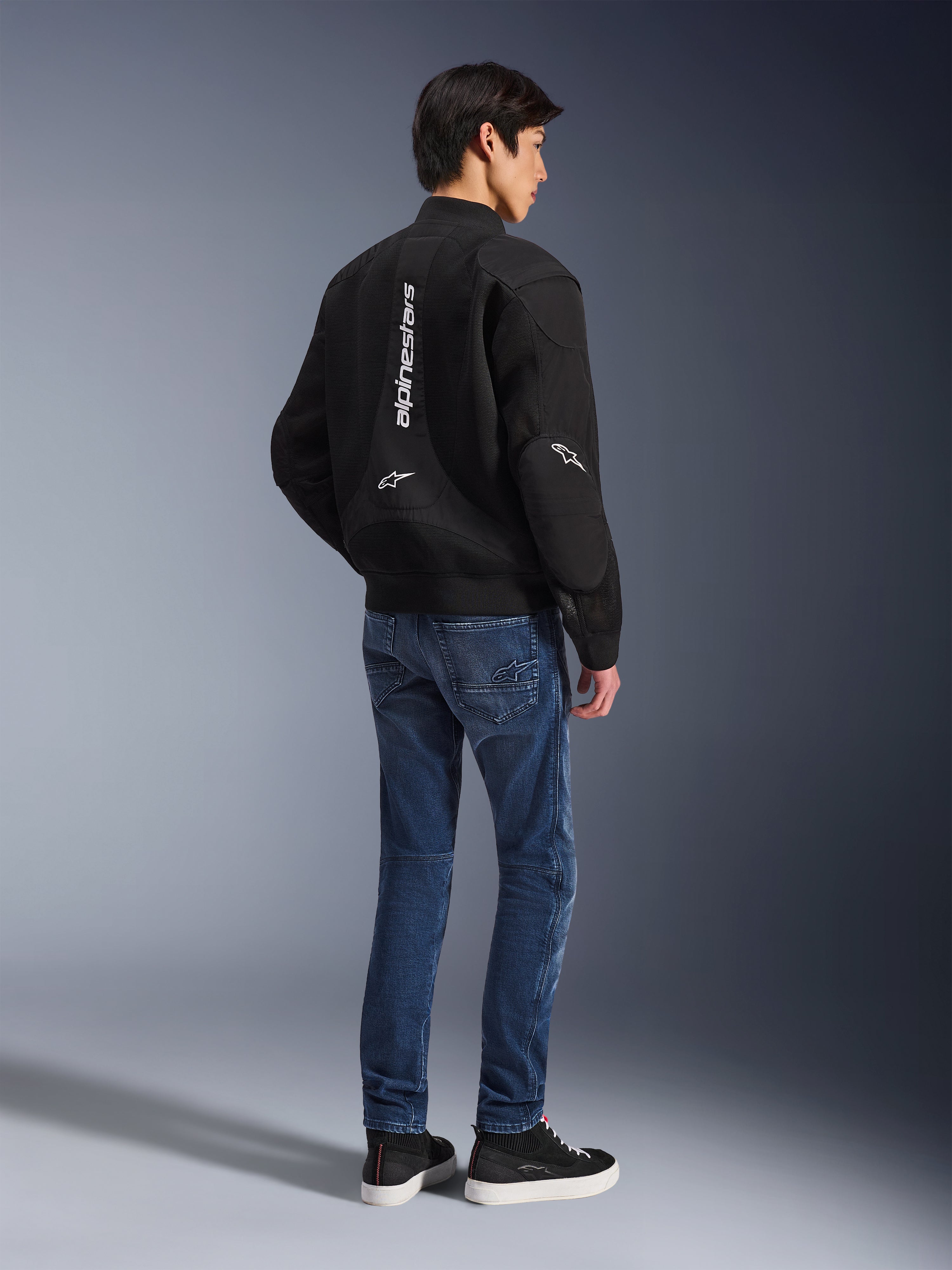 Modelo con la chaqueta Alpinestars Flight Air en color negro, vista desde atrás, combinada con vaqueros de moto azules y zapatillas negras, con marca Alpinestars vertical en blanco y paneles de ventilación de malla 3D.