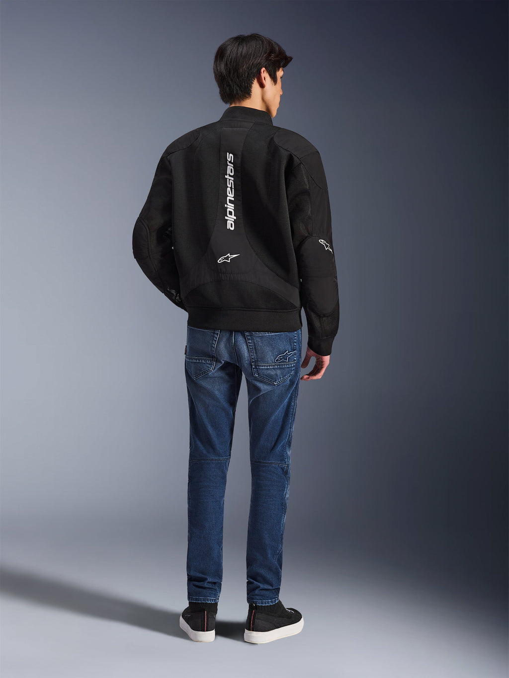 Modelo de espaldas con la Chaqueta Flight Air de Alpinestars, chaqueta de moto negra estilo bomber con paneles de malla 3D y logotipo blanco vertical, combinada con vaqueros de moto azules y zapatillas negras, equipamiento urbano contemporáneo para motociclista