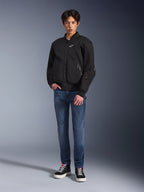 Modelo vistiendo la chaqueta Alpinestars Flight Air en color negro, una chaqueta de moto de estilo bomber urbano con paneles de malla 3D y cremallera frontal, combinada con pantalones vaqueros azules y zapatillas negras, equipamiento de conducción contemporáneo de inspiración urbana.