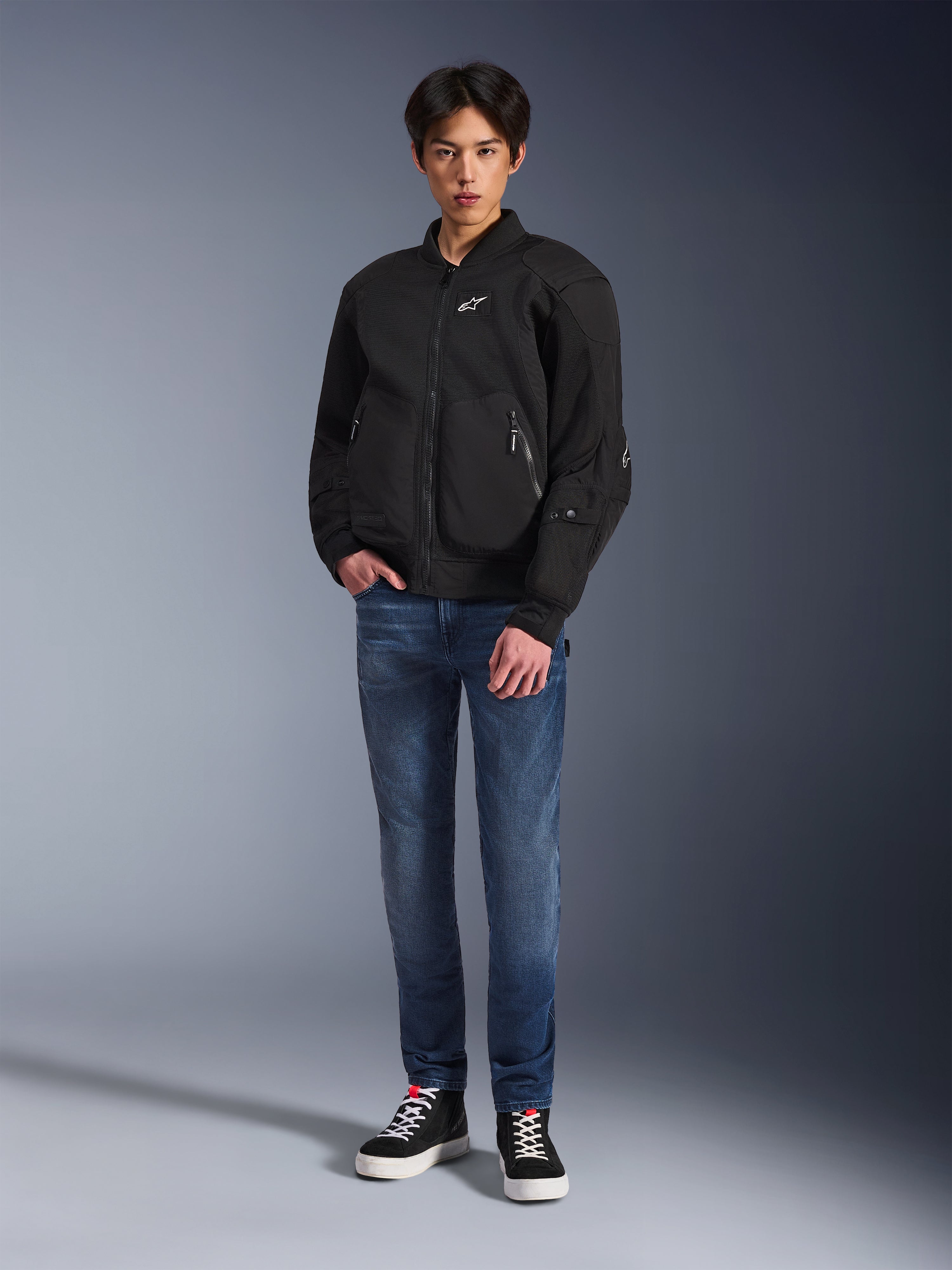 Modelo vistiendo la chaqueta Alpinestars Flight Air en color negro, una chaqueta de moto de estilo bomber urbano con paneles de malla 3D y cremallera frontal, combinada con pantalones vaqueros azules y zapatillas negras, equipamiento de conducción contemporáneo de inspiración urbana.