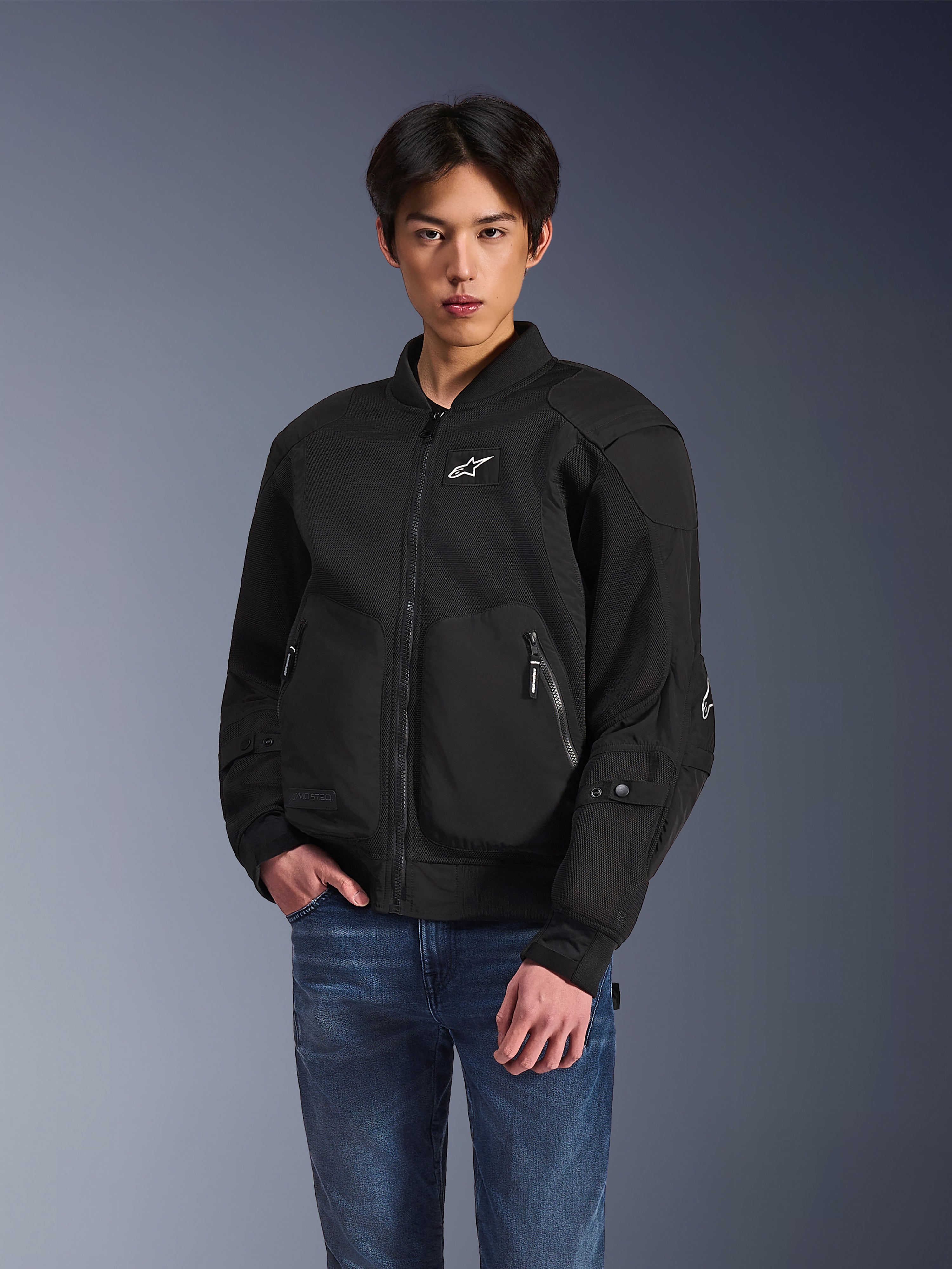 Chaqueta Alpinestars Flight Air, chaqueta de moto urbana negra, usada por un hombre con vaqueros azules, con una estética tipo bomber con paneles de malla, cremallera frontal y logotipo blanco en el pecho, diseñada para la conducción urbana en climas cálidos