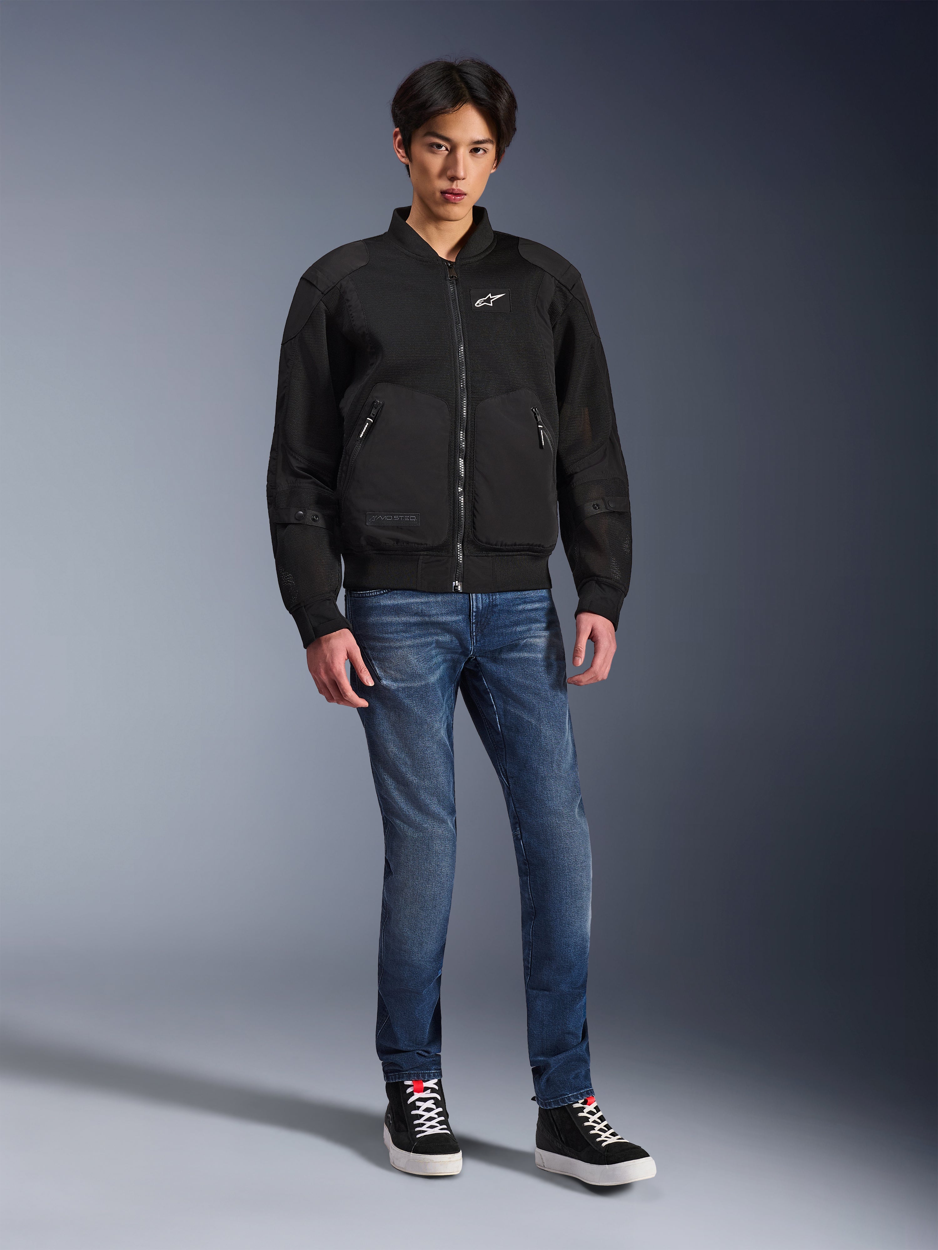 Chaqueta Alpinestars Flight Air, negra, chaqueta de moto estilo bomber de inspiración urbana con paneles de malla 3D y logotipo blanco en el pecho, usada por un modelo con vaqueros azules y zapatillas negras, indumentaria de conducción contemporánea