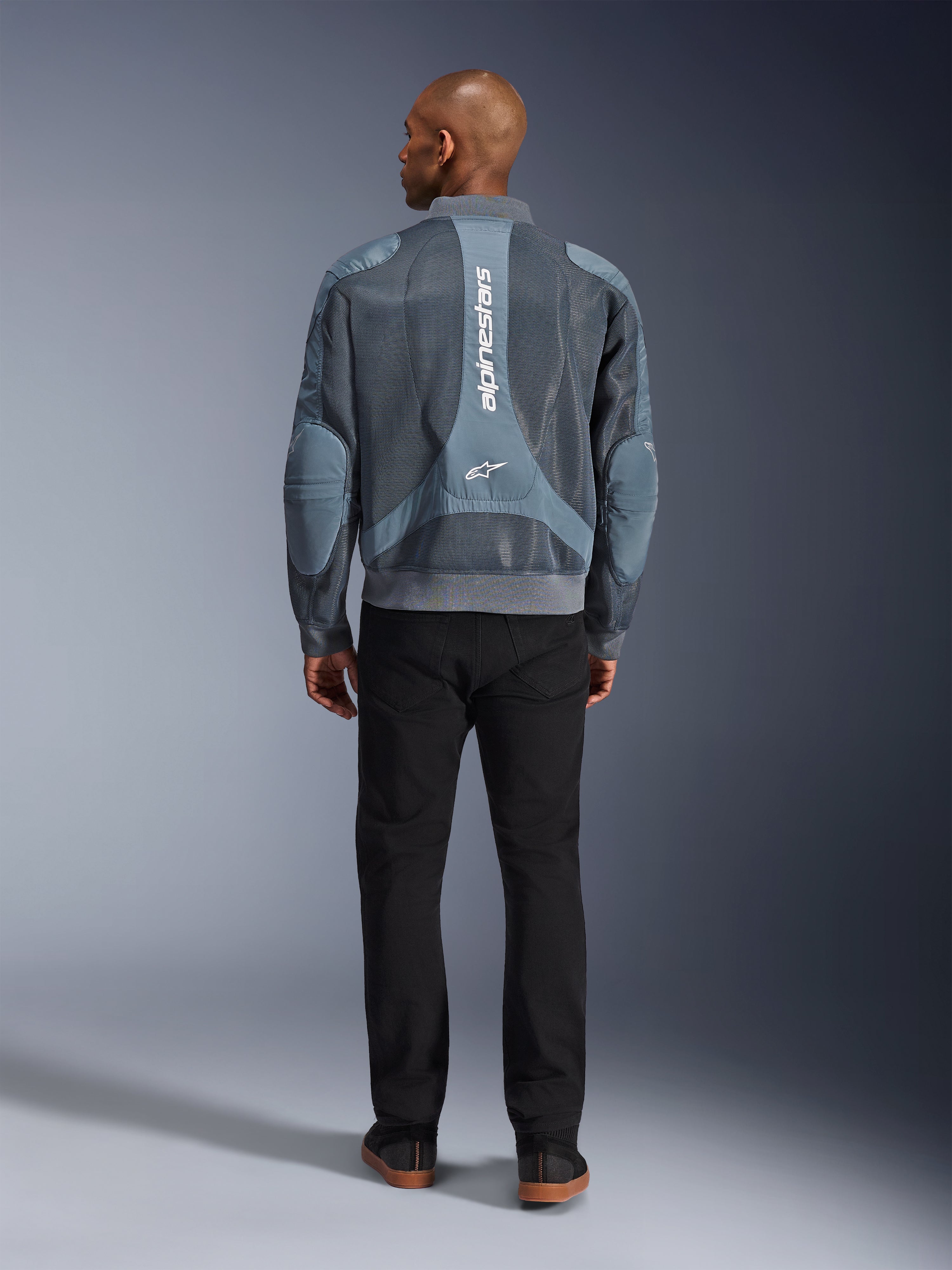 Alpinestars Flight Air Jacket en Storm Gray, vista trasera de un modelo masculino vistiendo una chaqueta de moto de estilo urbano con paneles de malla 3D y la marca Alpinestars en vertical, combinada con pantalones negros para un look urbano moderno