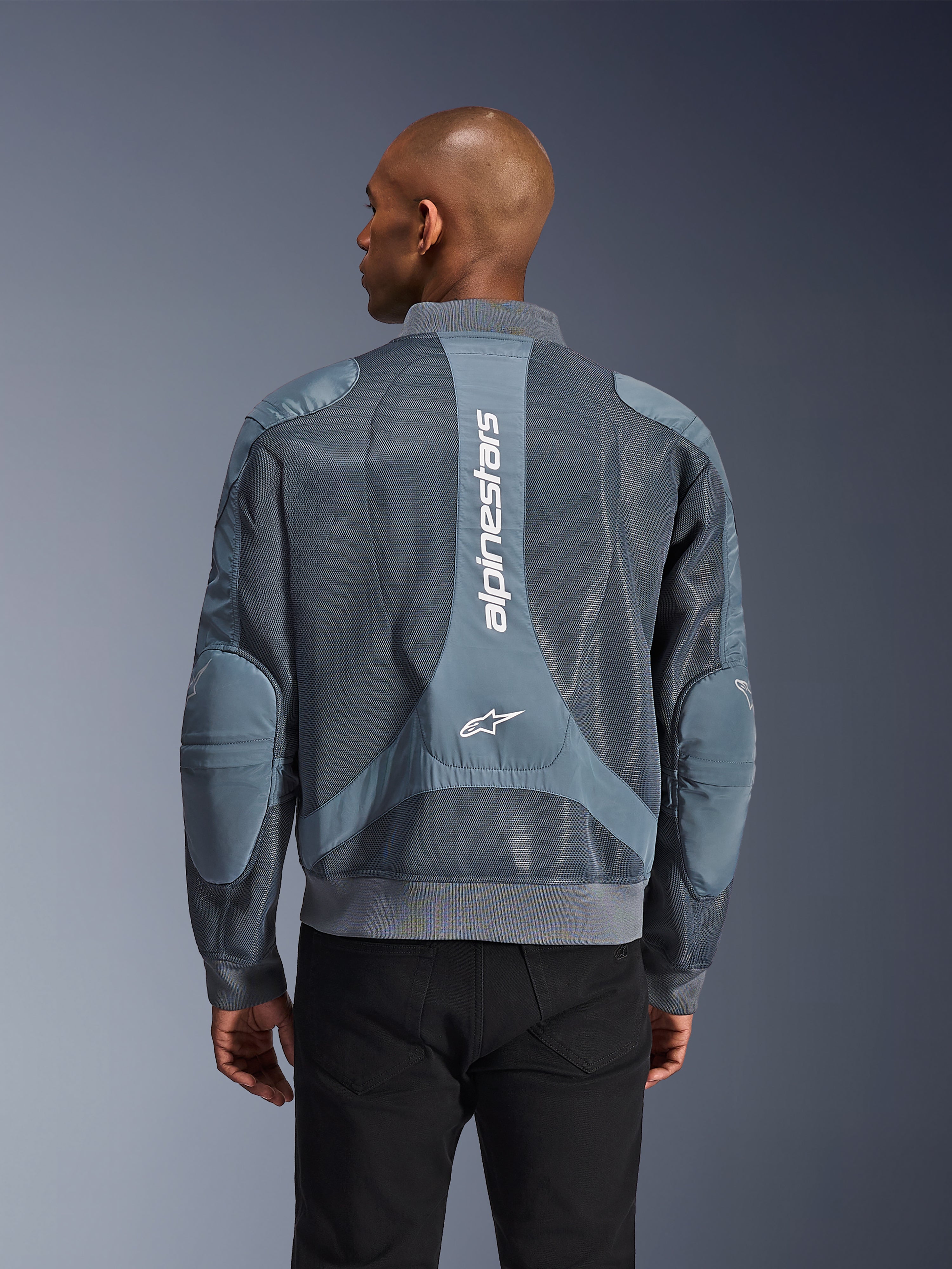 Chaqueta Alpinestars Flight Air en color Storm Gray, vista trasera en un modelo, chaqueta de moto urbana con paneles de malla 3D extendidos, marca Alpinestars vertical en la espalda y cuello y dobladillo acanalados estilo bomber.
