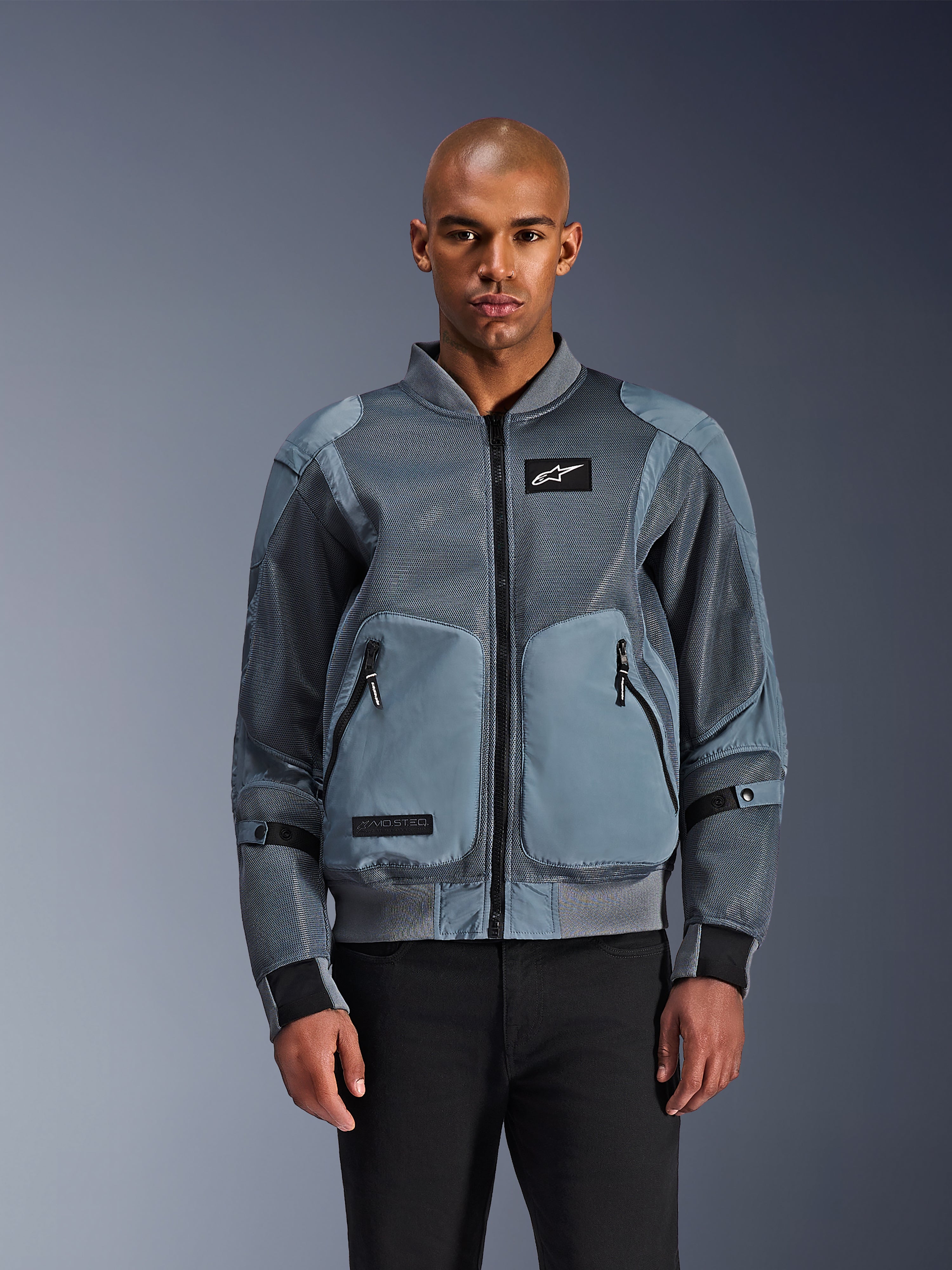 Chaqueta Alpinestars Flight Air, chaqueta de moto urbana estilo bomber, color Storm Gray, con paneles de ventilación de malla 3D y parche con el logotipo de Astars en blanco, vestida por un modelo masculino con pantalones negros, equipamiento de moto urbano contemporáneo