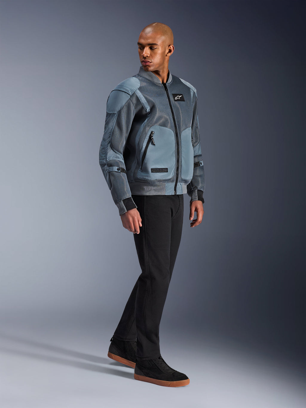Chaqueta Alpinestars Flight Air, Gris Storm Gray, chaqueta de motociclismo de inspiración bomber urbana, vestida por un modelo masculino con pantalones negros y zapatillas, con paneles de ventilación de malla 3D extendidos y bolsillos externos para protectores, equipamiento de estilo urbano para conducir en climas cálidos
