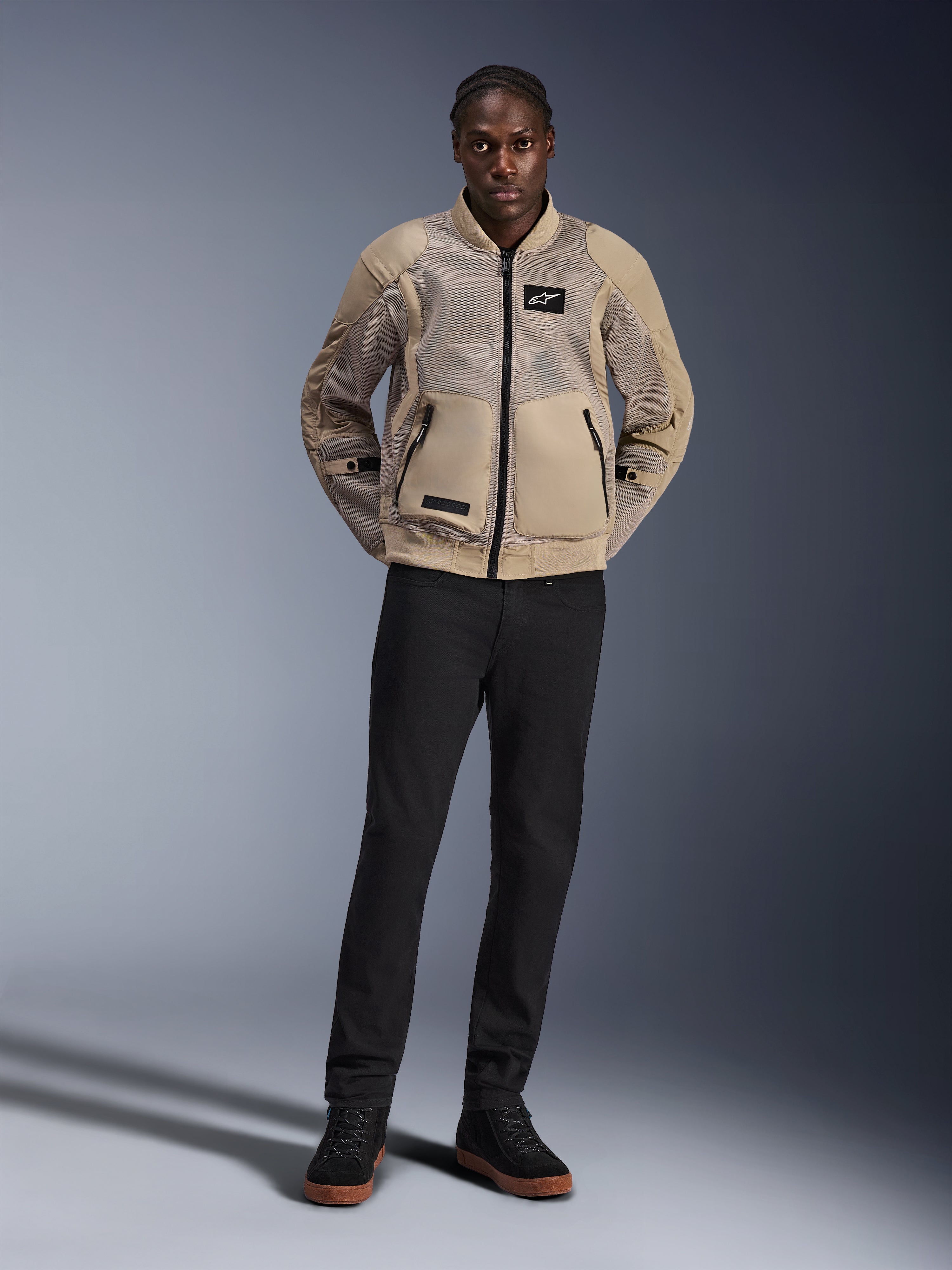 Modelo luciendo la chaqueta Alpinestars Flight Air en color marrón, chaqueta urbana para motociclista con paneles de malla 3D extendidos, cuello y puños acanalados de estilo bomber, combinada con pantalones negros y zapatos de conducción negros, equipamiento urbano contemporáneo