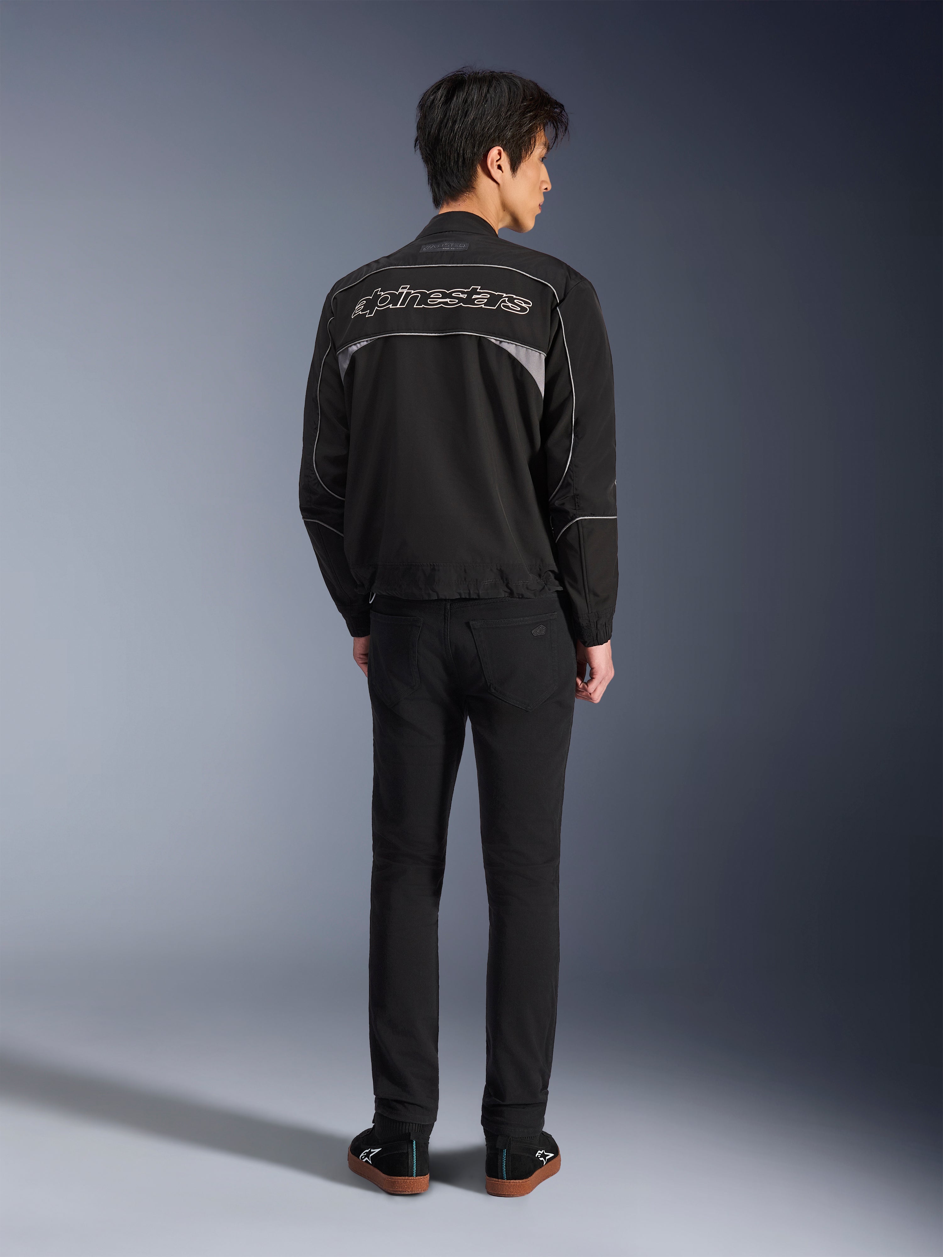 Alpinestars Unite Jacket, Black and Dark Gray motorcycle jacket, rear view showing Alpinestars outline logo on the back, gray reflective panels, minimalist urban design, worn with black pants and black shoes.