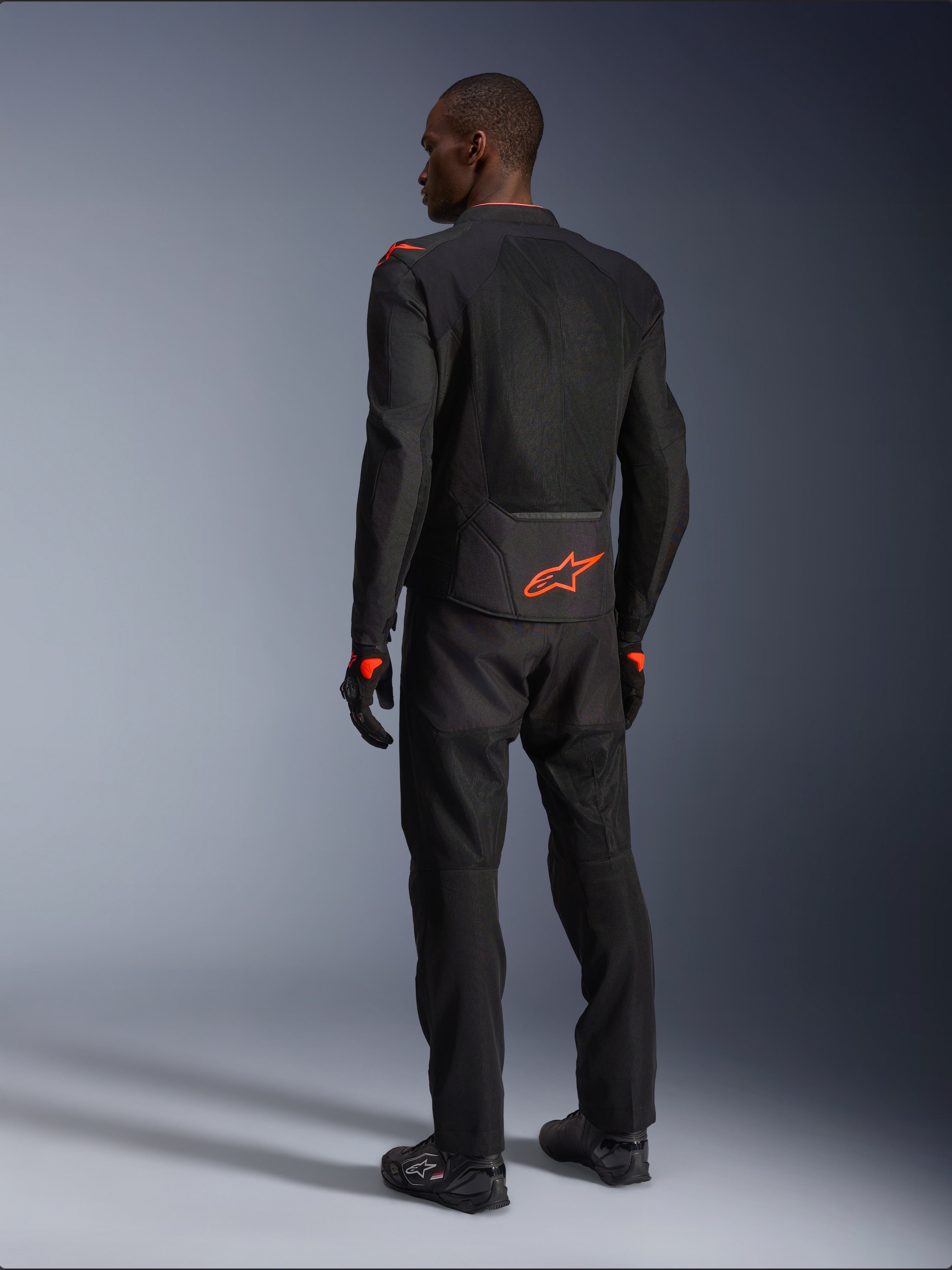Male rider from the back wearing the Alpinestars Viper Air V4 Jacket, Black and Red Fluo, featuring an orange Astars logo on the lower back and shoulders, mesh textile construction, styled with matching black motorcycle pants and boots.