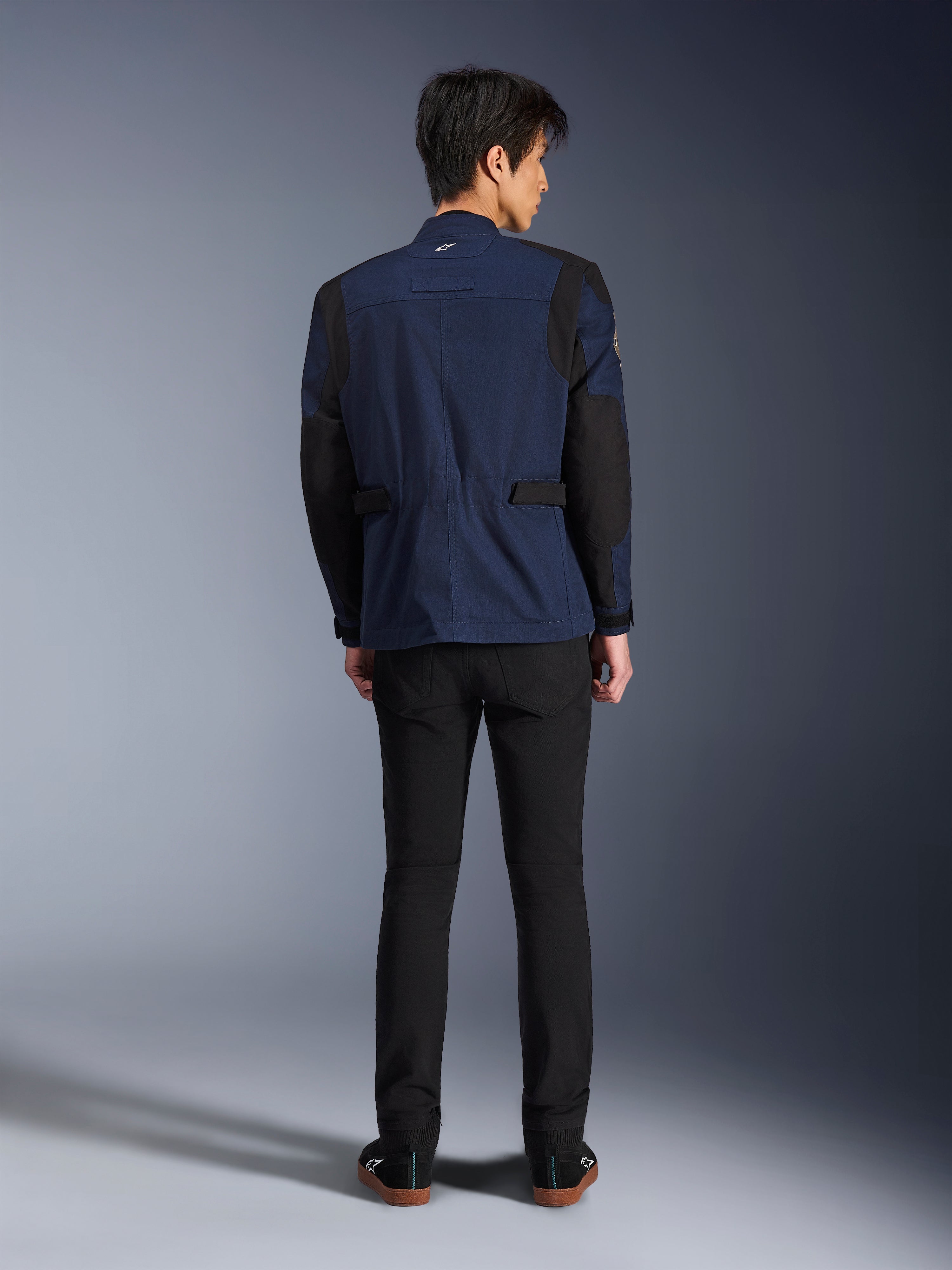 Model wearing the Alpinestars Dice Jacket in Dark Blue and Black, back view, urban motorcycle field jacket featuring box-pleated shoulders and adjustable waist tabs, paired with black pants and black sneakers.