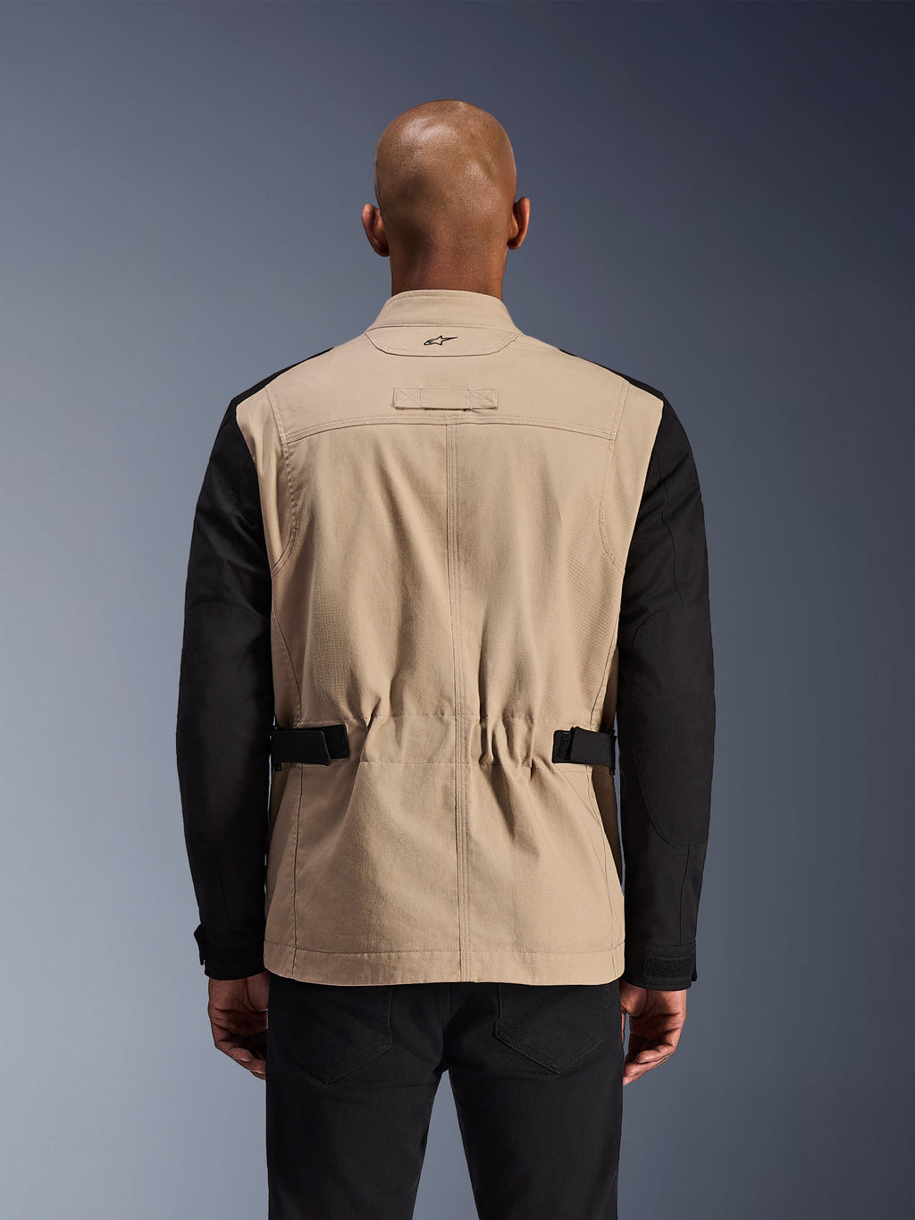 Chaqueta Alpinestars Dice, chaqueta de moto urbana, aluminio y negro, vista trasera en un modelo, con hombros plisados para movilidad, correas de cintura ajustables y construcción robusta de sarga de policotón.