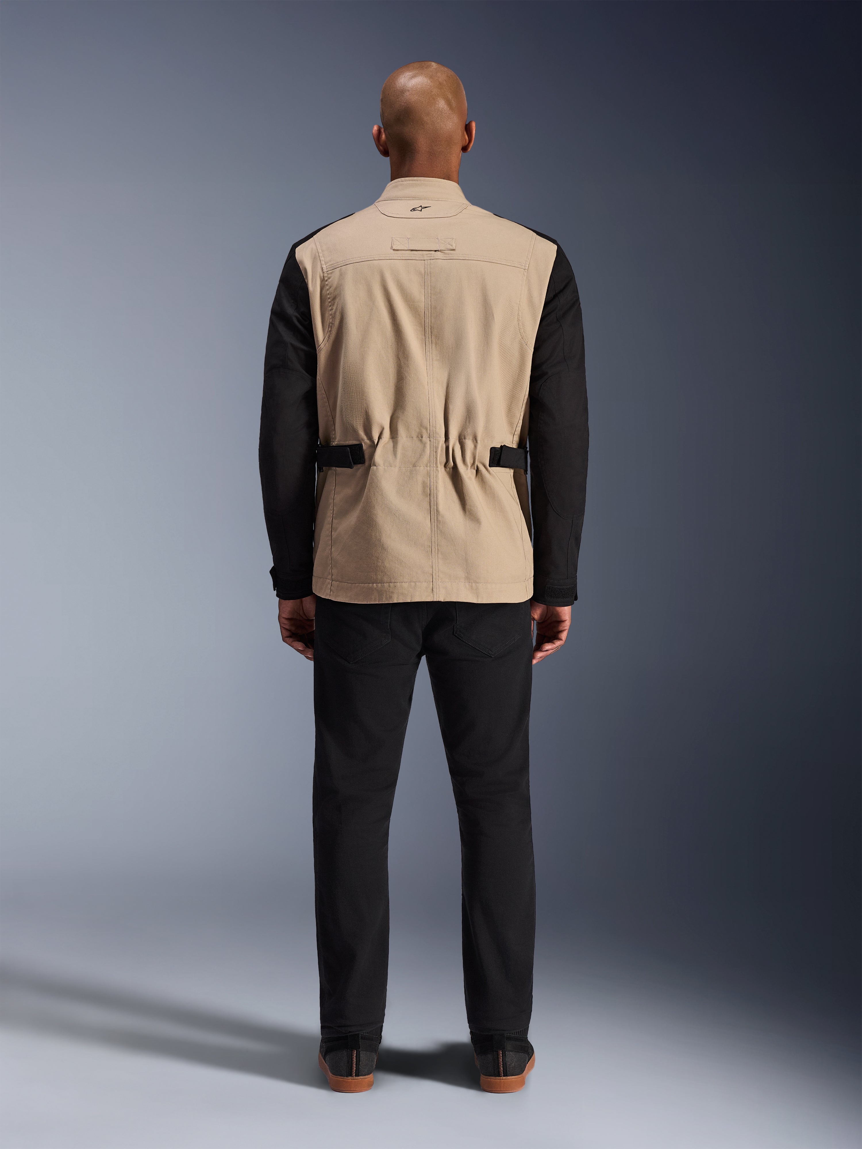 Chaqueta Dice de Alpinestars, chaqueta urbana para moto, beige y negro, vista posterior en modelo, con confección en sarga de policotón, correas de ajuste en la cintura y un pequeño logotipo bordado en el cuello, combinada con pantalones negros