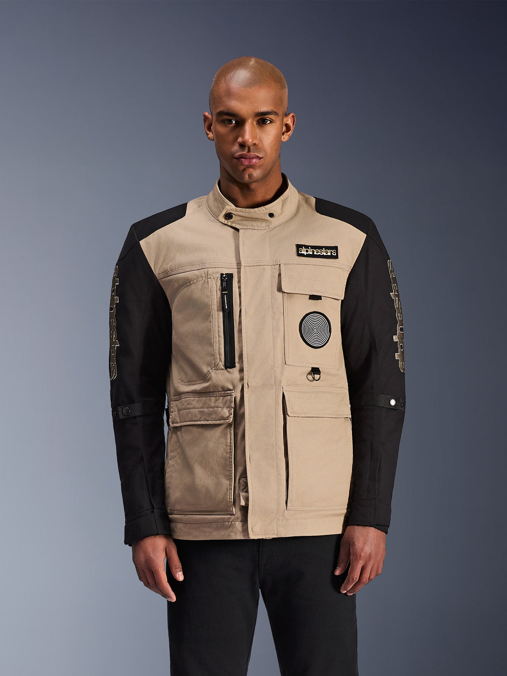 Alpinestars Dice Jacket, chaqueta de moto urbana, beige y negra, estética robusta de ropa de trabajo con múltiples bolsillos utilitarios y parches en el pecho, cuello alto con cierre de botón, logotipos llamativos en las mangas, usada por un modelo masculino