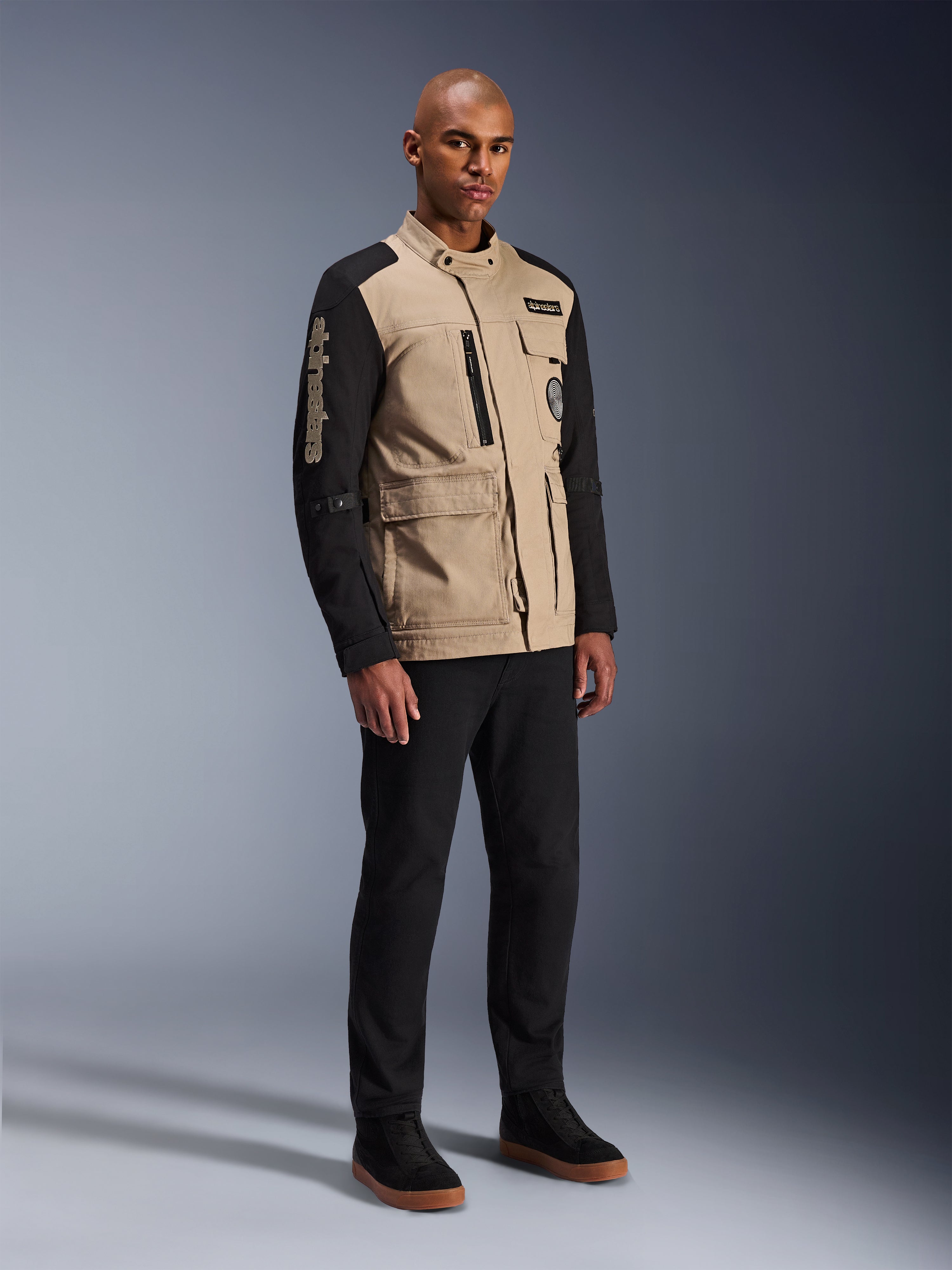 Chaqueta Alpinestars Dice, chaqueta urbana de motociclismo, Aluminum Black y tostado, diseño en dos tonos con mangas negras y torso caqui, lucida por un modelo con pantalones negros y zapatillas negras, estética de inspiración 'workwear' con bolsillos funcionales y marca bordada en la manga.