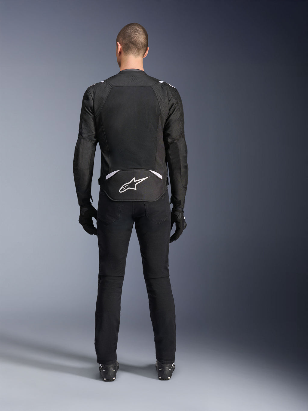 Chaqueta Alpinestars T-Sps Air V2, vista trasera, chaqueta de moto textil en negro y blanco, con un gran panel de malla en la espalda para un flujo de aire máximo y logotipo de Astars en blanco en la parte inferior de la espalda, combinada con pantalones y guantes de conducción negros