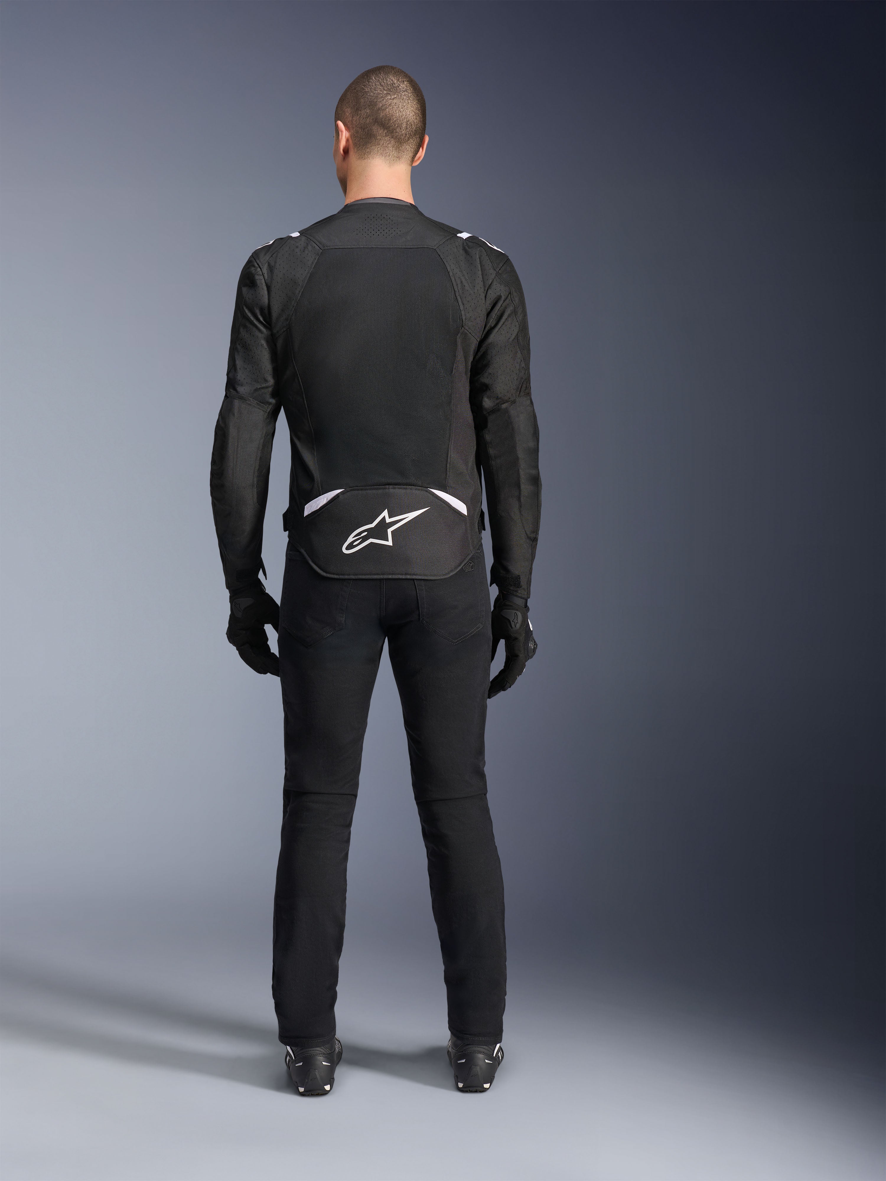 Chaqueta Alpinestars T-Sps Air V2, vista trasera, chaqueta de moto textil en negro y blanco, con un gran panel de malla en la espalda para un flujo de aire máximo y logotipo de Astars en blanco en la parte inferior de la espalda, combinada con pantalones y guantes de conducción negros