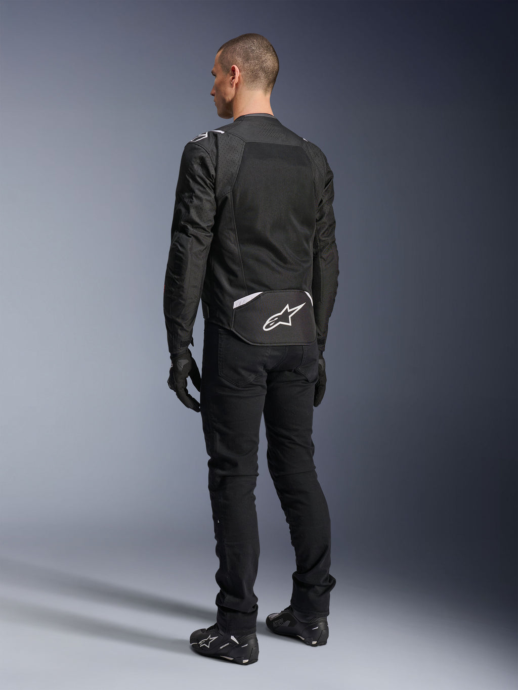 Chaqueta Alpinestars T-Sps Air V2, chaqueta de moto textil, negra, vista trasera que muestra un gran panel de ventilación de malla y el logotipo blanco de Alpinestars en la parte inferior de la espalda, combinada con pantalones y guantes negros para un estilo urbano deportivo.
