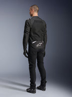 Chaqueta Alpinestars T-Sps Air V2, chaqueta de moto textil, negra, vista trasera que muestra un gran panel de ventilación de malla y el logotipo blanco de Alpinestars en la parte inferior de la espalda, combinada con pantalones y guantes negros para un estilo urbano deportivo.