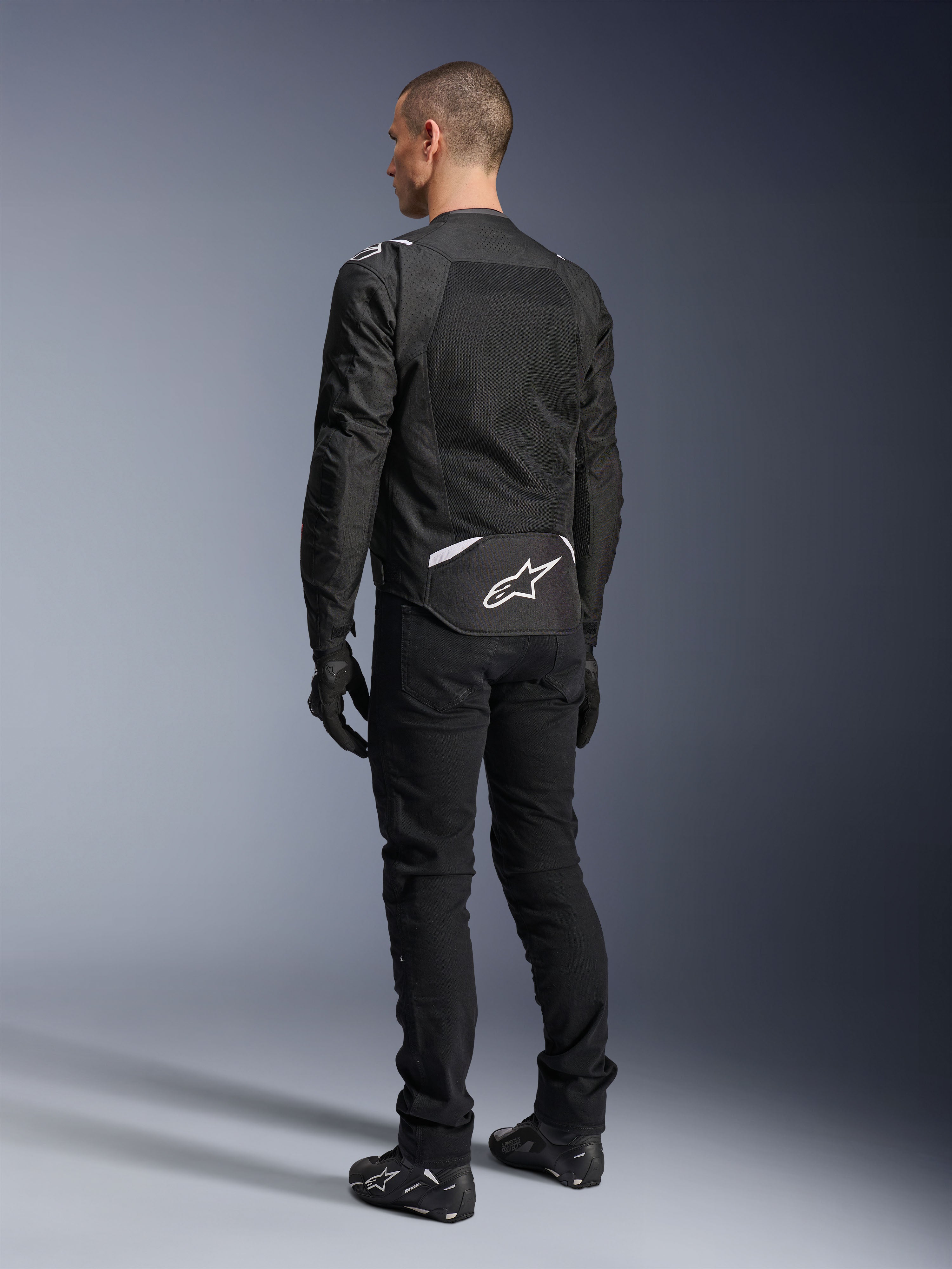 Chaqueta Alpinestars T-Sps Air V2, chaqueta de moto textil, negra, vista trasera que muestra un gran panel de ventilación de malla y el logotipo blanco de Alpinestars en la parte inferior de la espalda, combinada con pantalones y guantes negros para un estilo urbano deportivo.