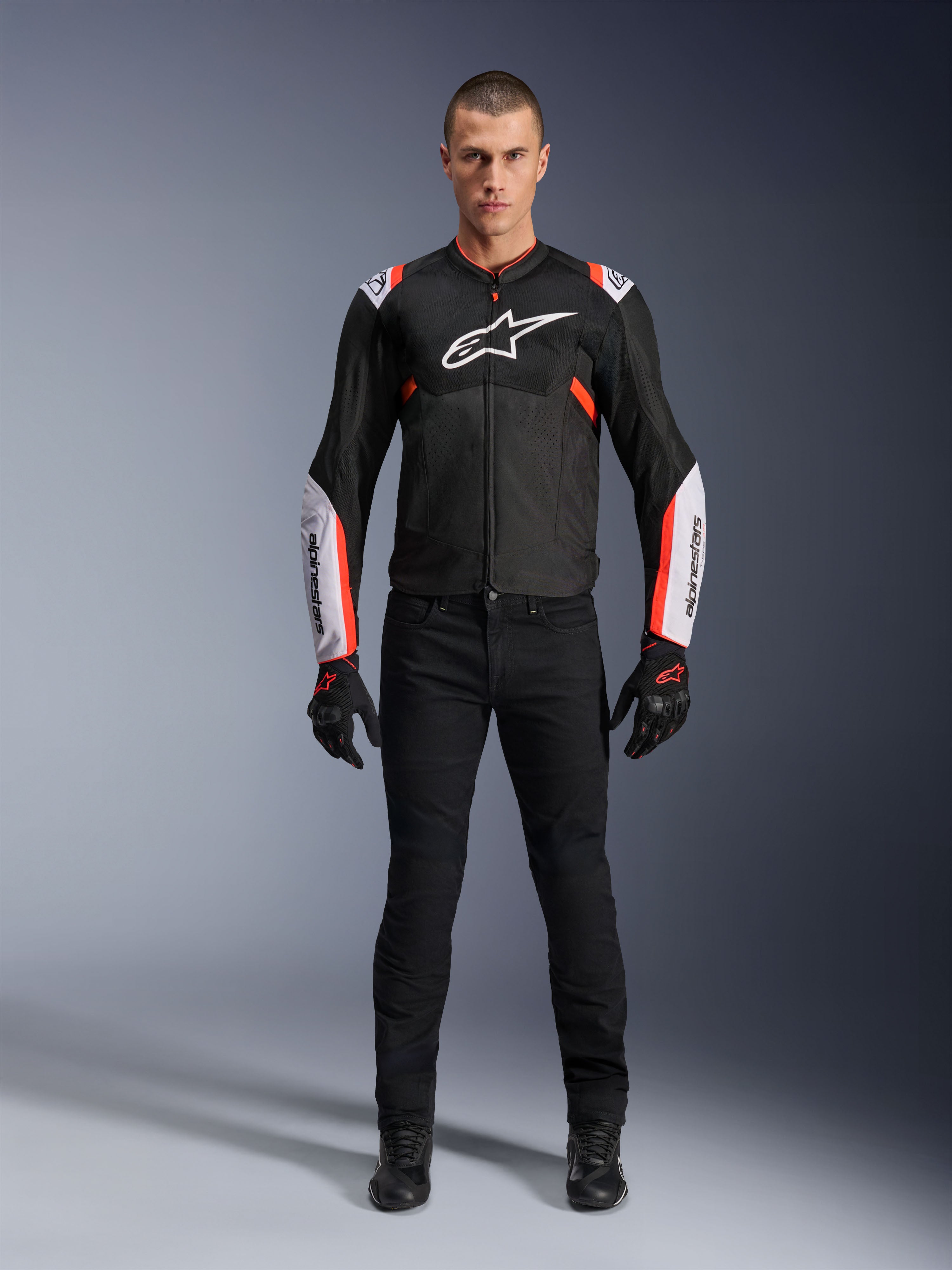 Modelo masculino con la chaqueta Alpinestars T-Sps Air V2 en negro, blanco y rojo flúor, con grandes paneles de malla técnica y logotipos blancos llamativos en el pecho y las mangas, combinada con pantalones técnicos de conducción y guantes negros, equipamiento deportivo urbano para moto