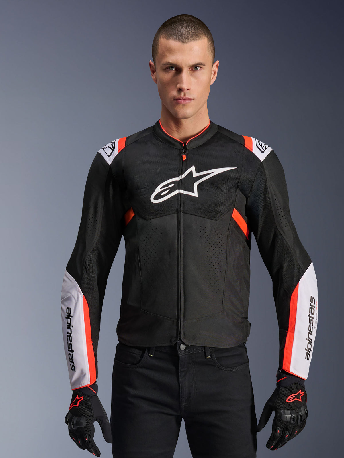 Chaqueta Alpinestars T-Sps Air V2, negra con detalles en blanco y rojo flúor, chaqueta deportiva de moto para hombre con diseño de ventilación de malla, logotipo grande en el pecho y mangas con la marca, combinada con guantes y pantalones negros.