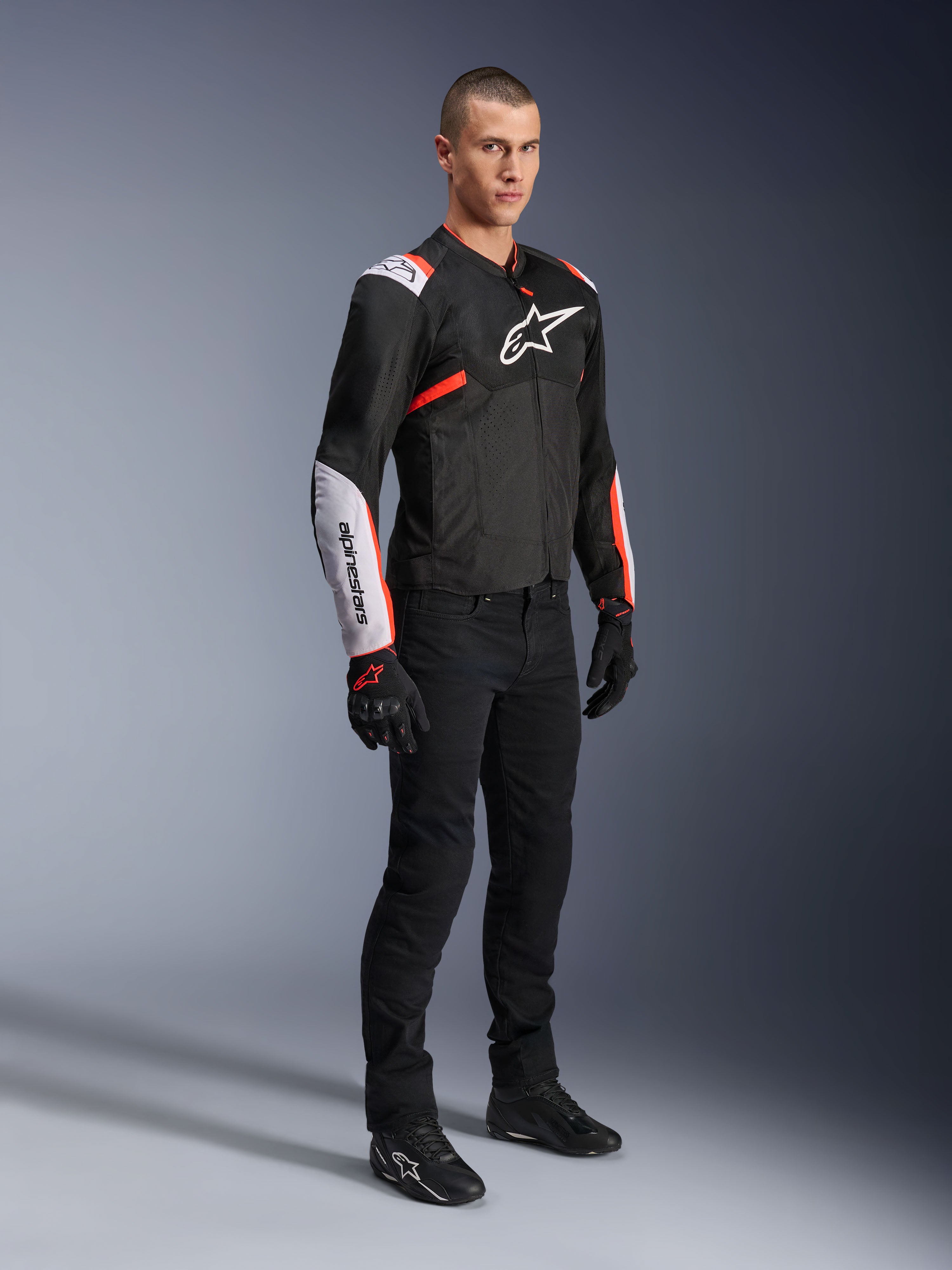 Chaqueta Alpinestars T-Sps Air V2, negro, blanco y rojo flúor, vestida por un modelo masculino con pantalones y guantes de moto negros, chaqueta textil de moto con amplios paneles de malla, secciones blancas en hombros y antebrazos, y un logotipo de Alpinestars destacado en el pecho.