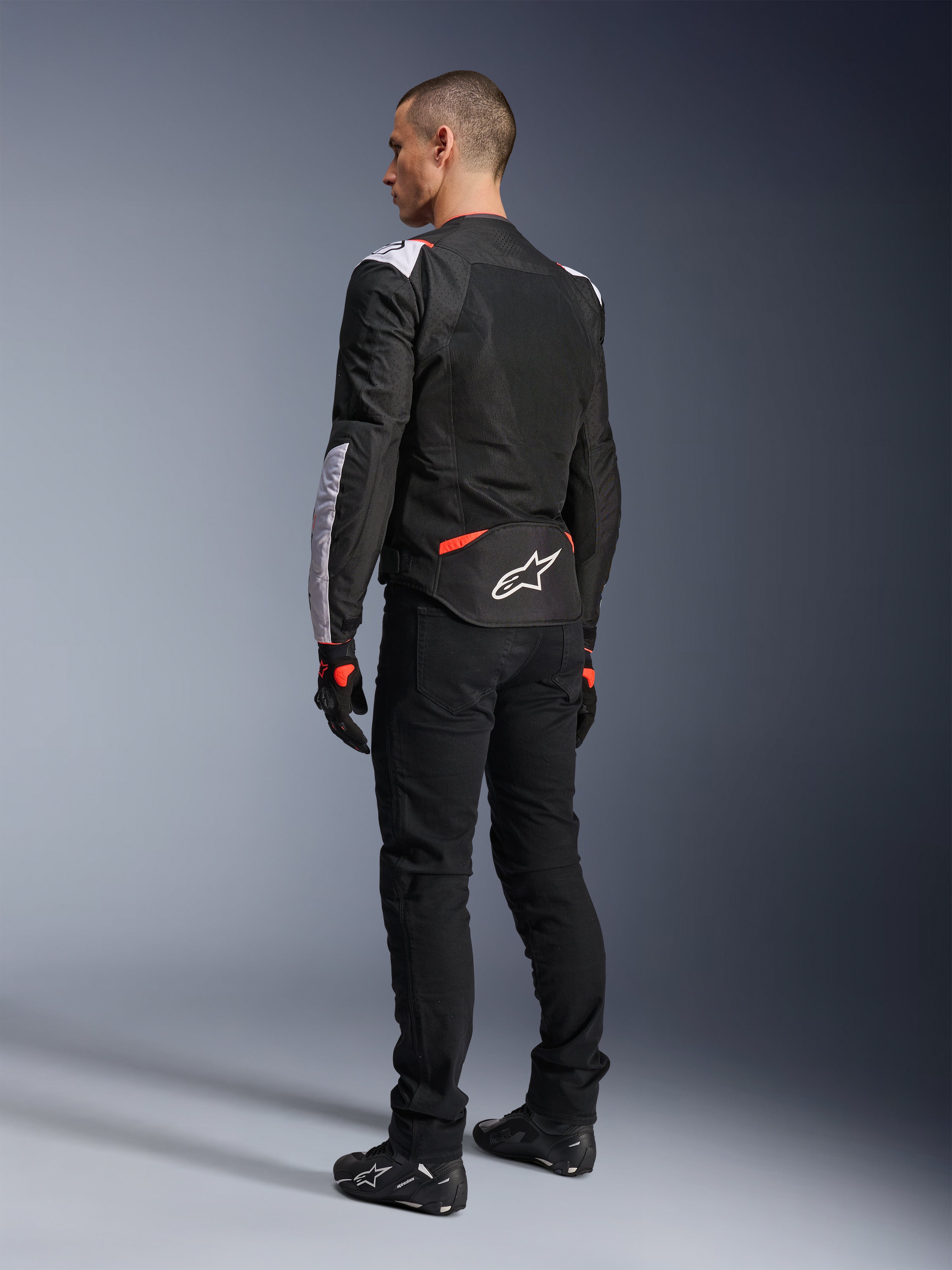 Chaqueta Alpinestars T-Sps Air V2, chaqueta textil de moto, negra, blanca y roja flúor, vista trasera en modelo, paneles de malla para ventilación, logotipo de Astars en blanco en la zona lumbar, combinada con pantalones de moto y guantes negros
