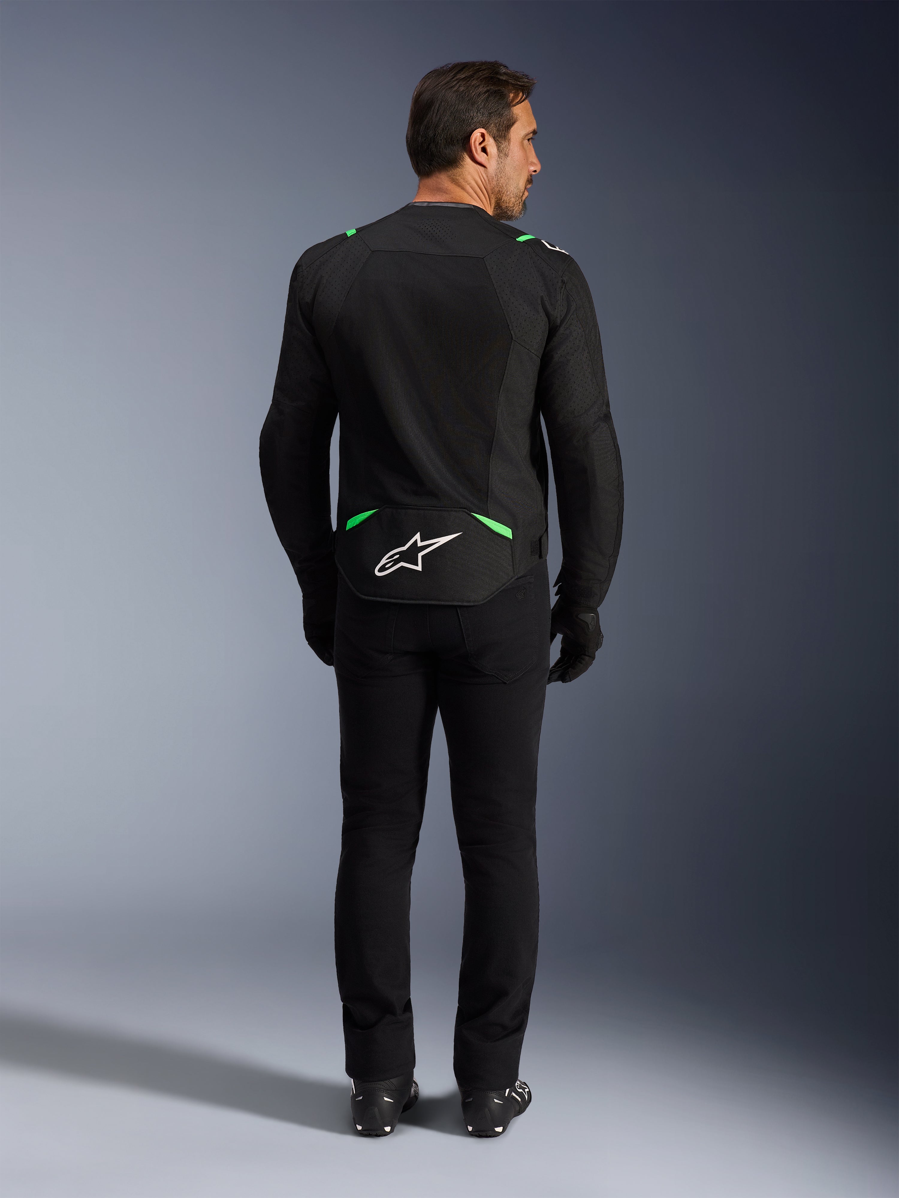 Chaqueta Alpinestars T-SPS Air V2, chaqueta de moto, negro y verde flúor, vista trasera con logo blanco en la parte inferior de la espalda, combinada con pantalones y botas negros, diseño de malla ventilada para conducción deportiva