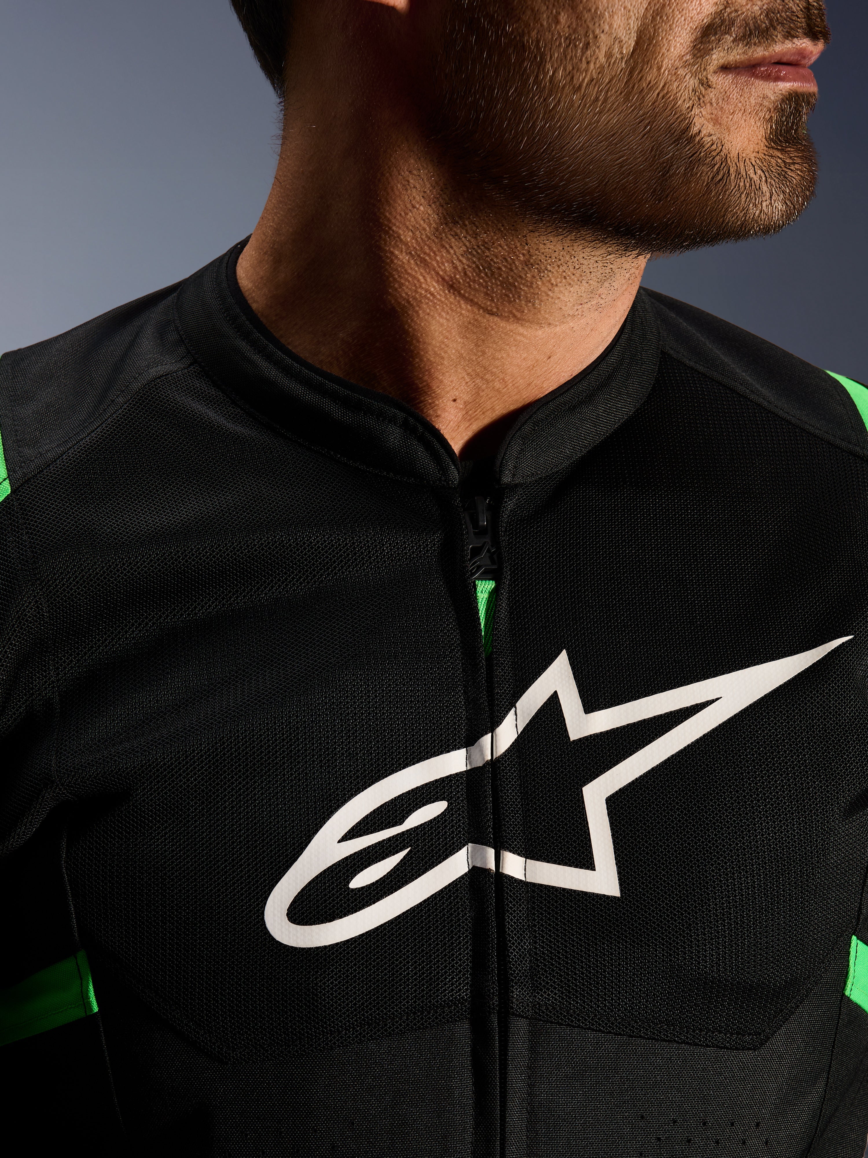 Primer plano de la chaqueta Alpinestars T-SPS Air V2, en color negro y verde flúor, con paneles de malla técnica transpirable, cremallera central y el destacado logotipo blanco de la estrella de Alpinestars en el pecho, equipamiento de motociclismo de alto rendimiento