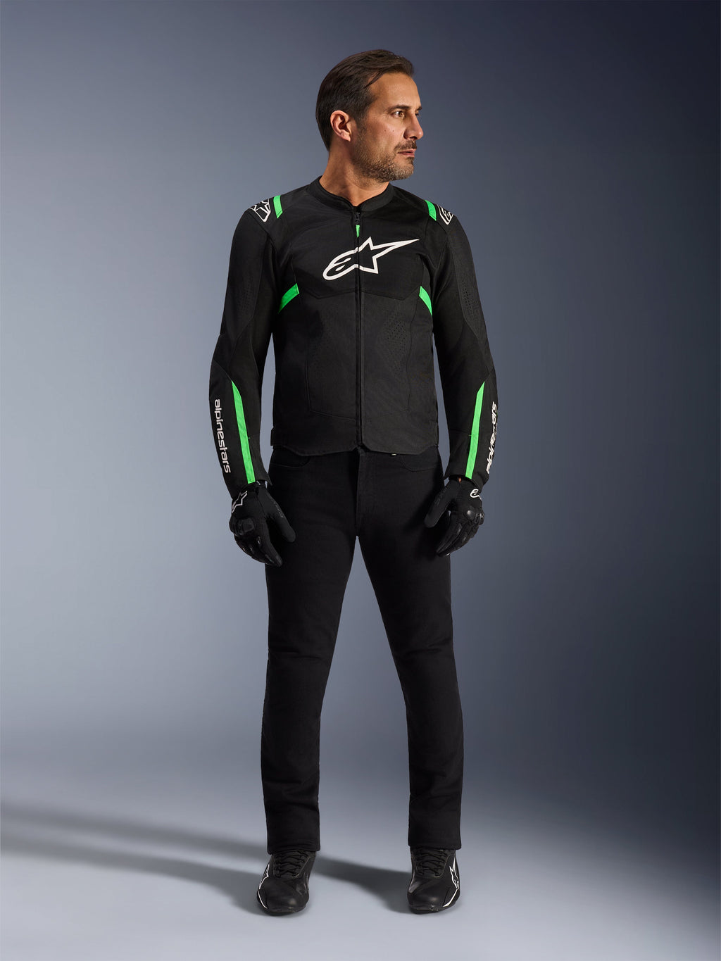 Hombre con la chaqueta Alpinestars T-SPS Air V2 en Black Fluo Green, combinada con pantalones negros, guantes y calzado de conducción, con un diseño de corte deportivo con logotipos blancos y paneles de malla para un estilo de conducción urbana moderno.