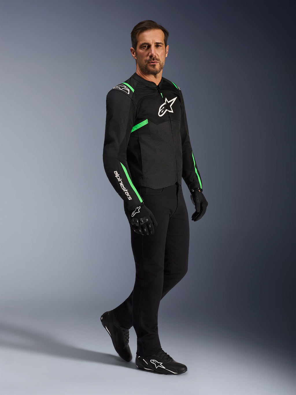 Modelo masculino luciendo la chaqueta Alpinestars T-SPS Air V2 en negro y verde flúor, combinada con pantalones de motociclismo y guantes negros, chaqueta textil de corte deportivo con paneles de malla y detalles neón, conjunto completo de equipamiento urbano para motorista