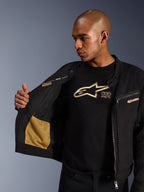 Alpinestars Bruiser Jacket, Black urban motorcycle jacket, close-up view showing open interior with gold mesh pocket and 'Alpinestars Protects' branding, worn over a black logo t-shirt, textile riding gear with workwear aesthetic