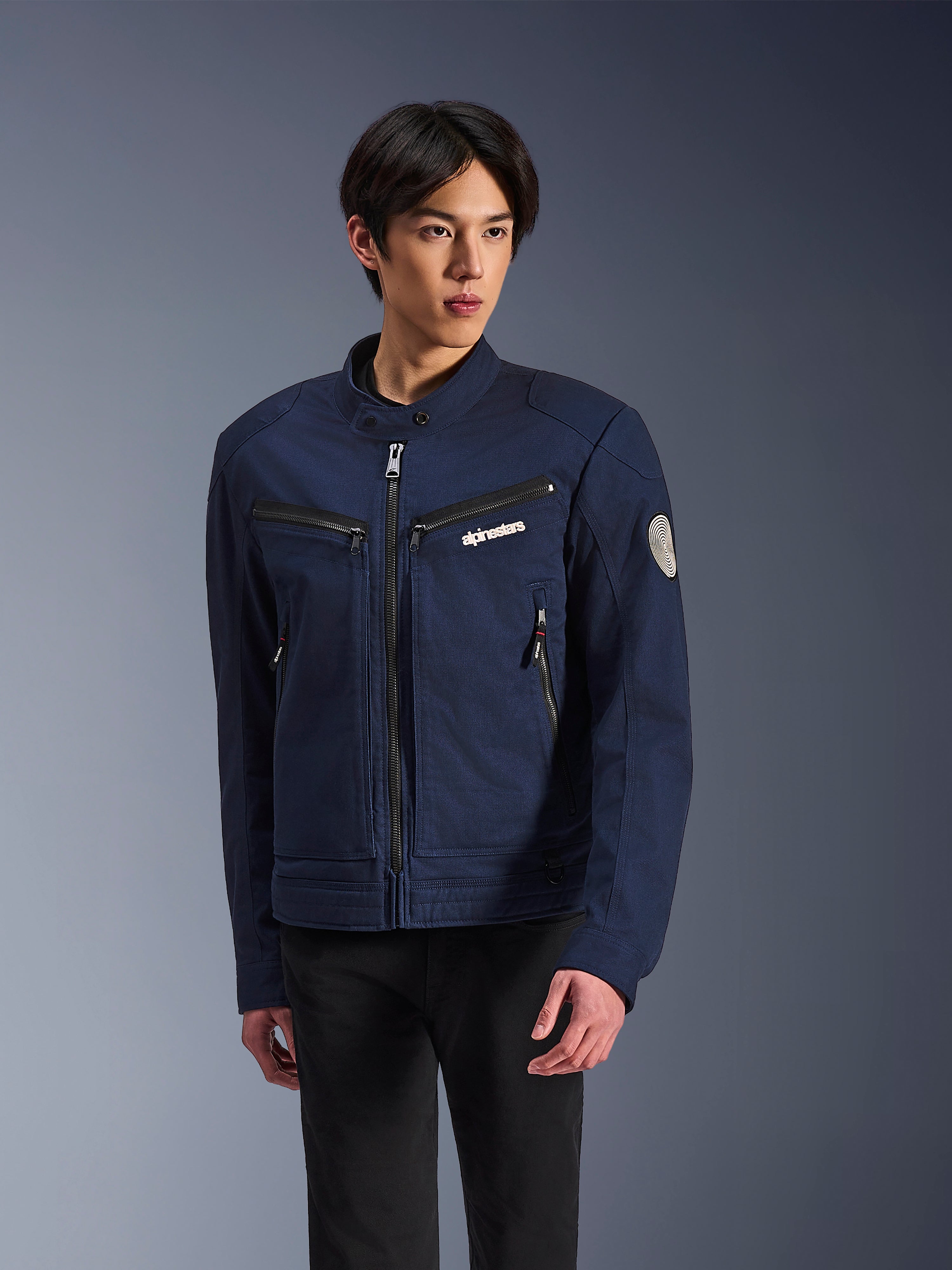 Chaqueta Alpinestars Bruiser, chaqueta textil de moto en azul oscuro, vestida por un hombre con pantalones negros, con bolsillos con cremallera, logotipo blanco de Alpinestars en el pecho y parche circular Oscar en la manga, estilo urbano para motoristas