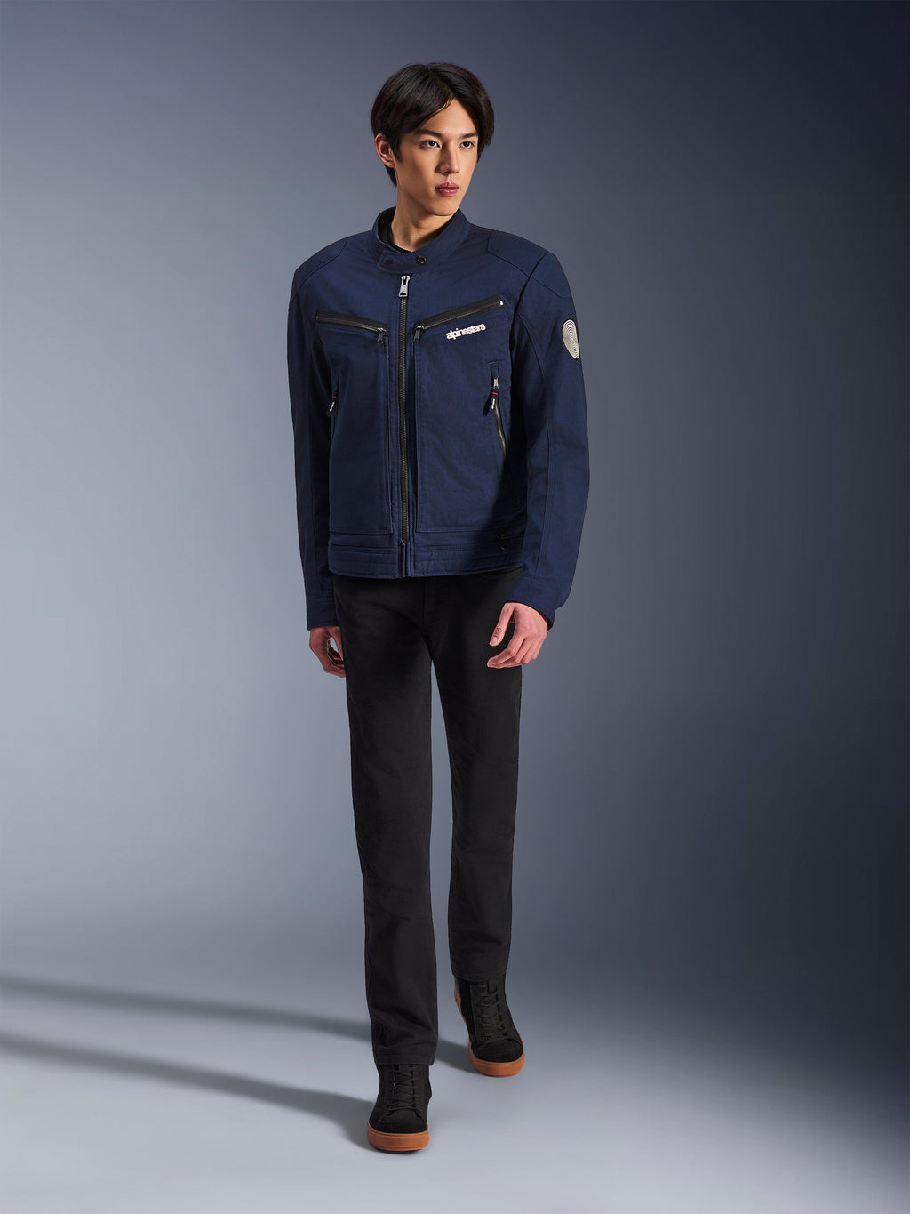 Modelo masculino vistiendo la chaqueta Alpinestars Bruiser en Dark Blue, chaqueta de moto textil urbana, combinada con pantalones negros y zapatillas negras, estética inspirada en la ropa de trabajo con bolsillos con cremallera y parche circular en la manga, estilo streetwear contemporáneo para montar en moto.