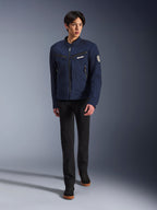 Modelo masculino vistiendo la chaqueta Alpinestars Bruiser en Dark Blue, chaqueta de moto textil urbana, combinada con pantalones negros y zapatillas negras, estética inspirada en la ropa de trabajo con bolsillos con cremallera y parche circular en la manga, estilo streetwear contemporáneo para montar en moto.