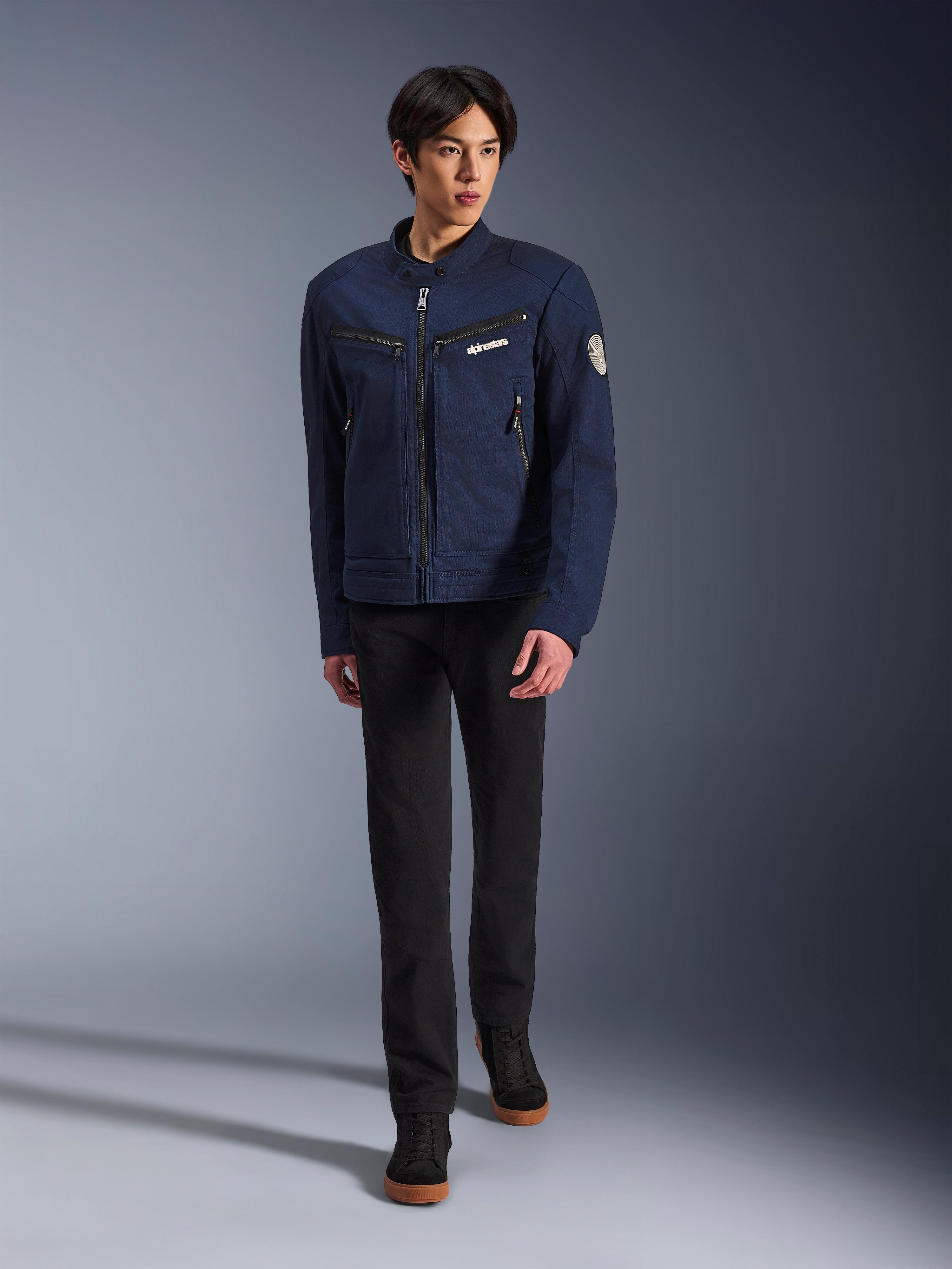 Modelo masculino vistiendo la chaqueta Alpinestars Bruiser en Dark Blue, chaqueta de moto textil urbana, combinada con pantalones negros y zapatillas negras, estética inspirada en la ropa de trabajo con bolsillos con cremallera y parche circular en la manga, estilo streetwear contemporáneo para montar en moto.