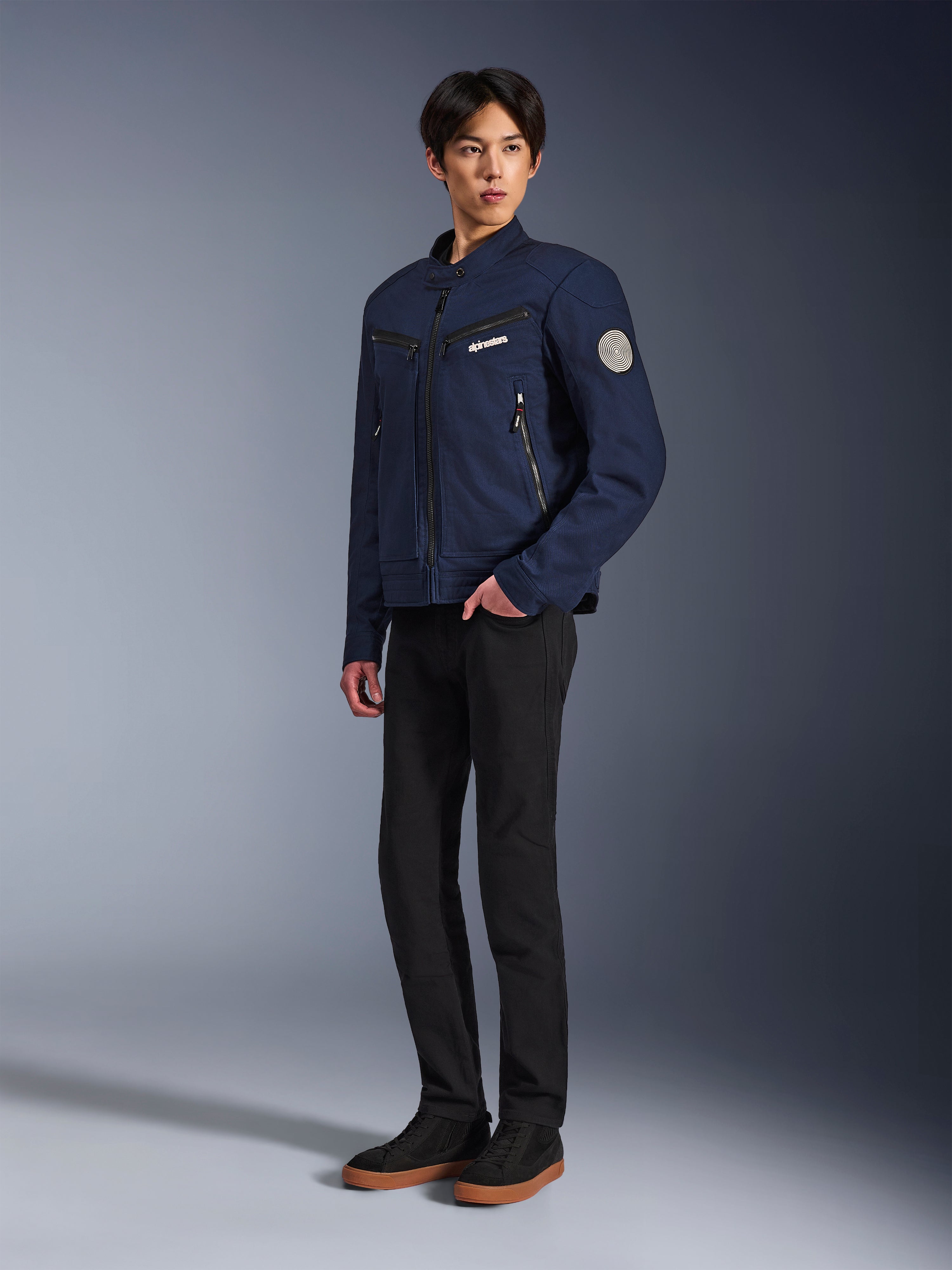 Chaqueta Alpinestars Bruiser en azul oscuro, chaqueta de moto urbana textil con diseño inspirado en la ropa de trabajo, con bolsillos con cremallera en el pecho y parche Oscar en la manga, usada con pantalones negros y zapatillas casuales.