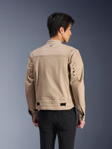 Chaqueta Alpinestars Bruiser, chaqueta de moto urbana marrón, vista trasera que muestra los hombros plisados articulados y las correas de ajuste en la cintura, usada con pantalones negros, equipamiento de conducción contemporáneo inspirado en la ropa de trabajo