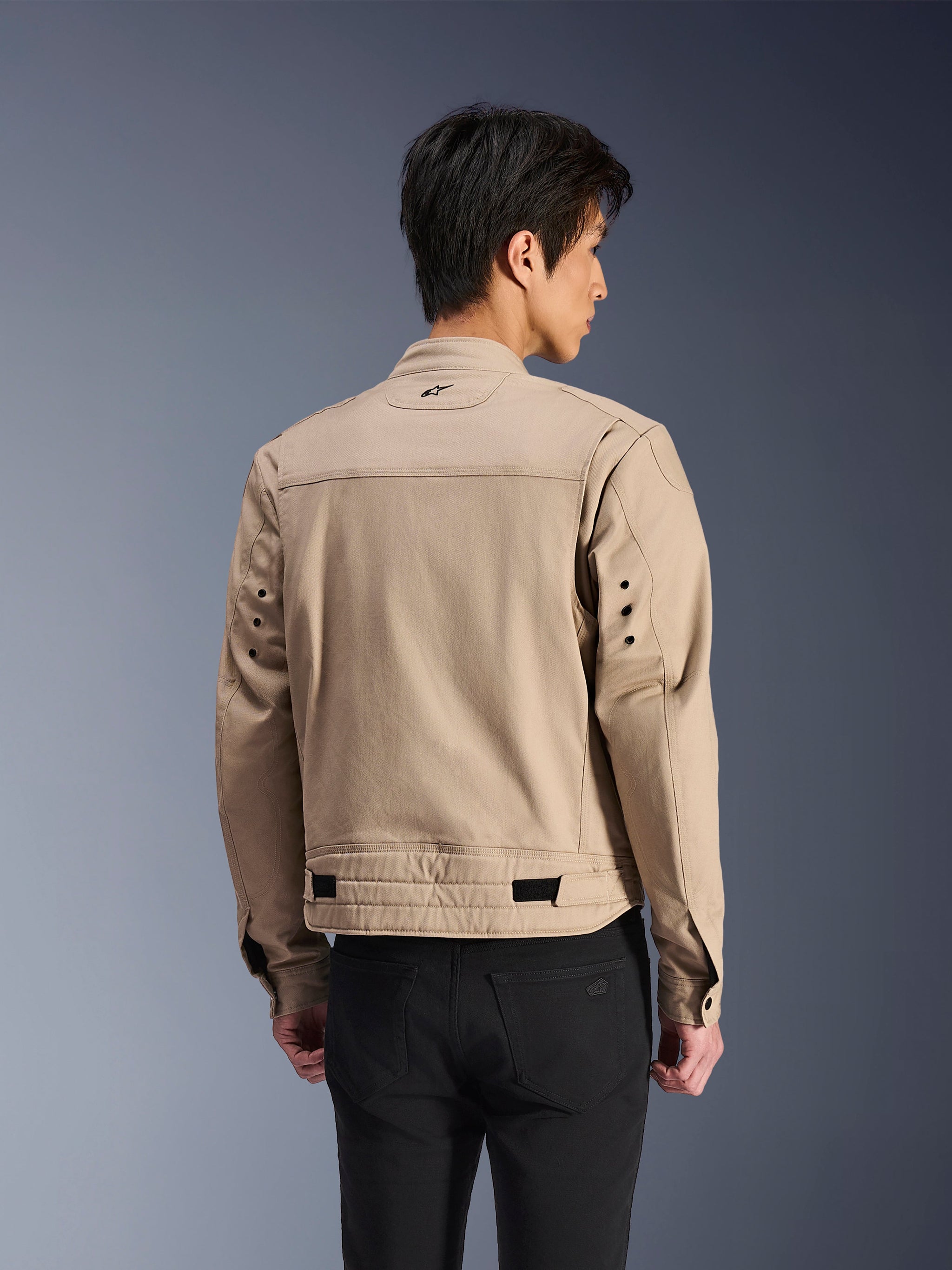 Chaqueta Alpinestars Bruiser, chaqueta de moto urbana marrón, vista trasera que muestra los hombros plisados articulados y las correas de ajuste en la cintura, usada con pantalones negros, equipamiento de conducción contemporáneo inspirado en la ropa de trabajo