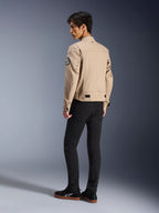 Modelo masculino de espaldas vistiendo la chaqueta Alpinestars Bruiser en color tostado, chaqueta de moto urbana contemporánea con estilo de ropa de trabajo, con un parche circular en la manga, correas de ajuste en la cintura y hombros articulados, combinada con pantalones negros y zapatillas negras.