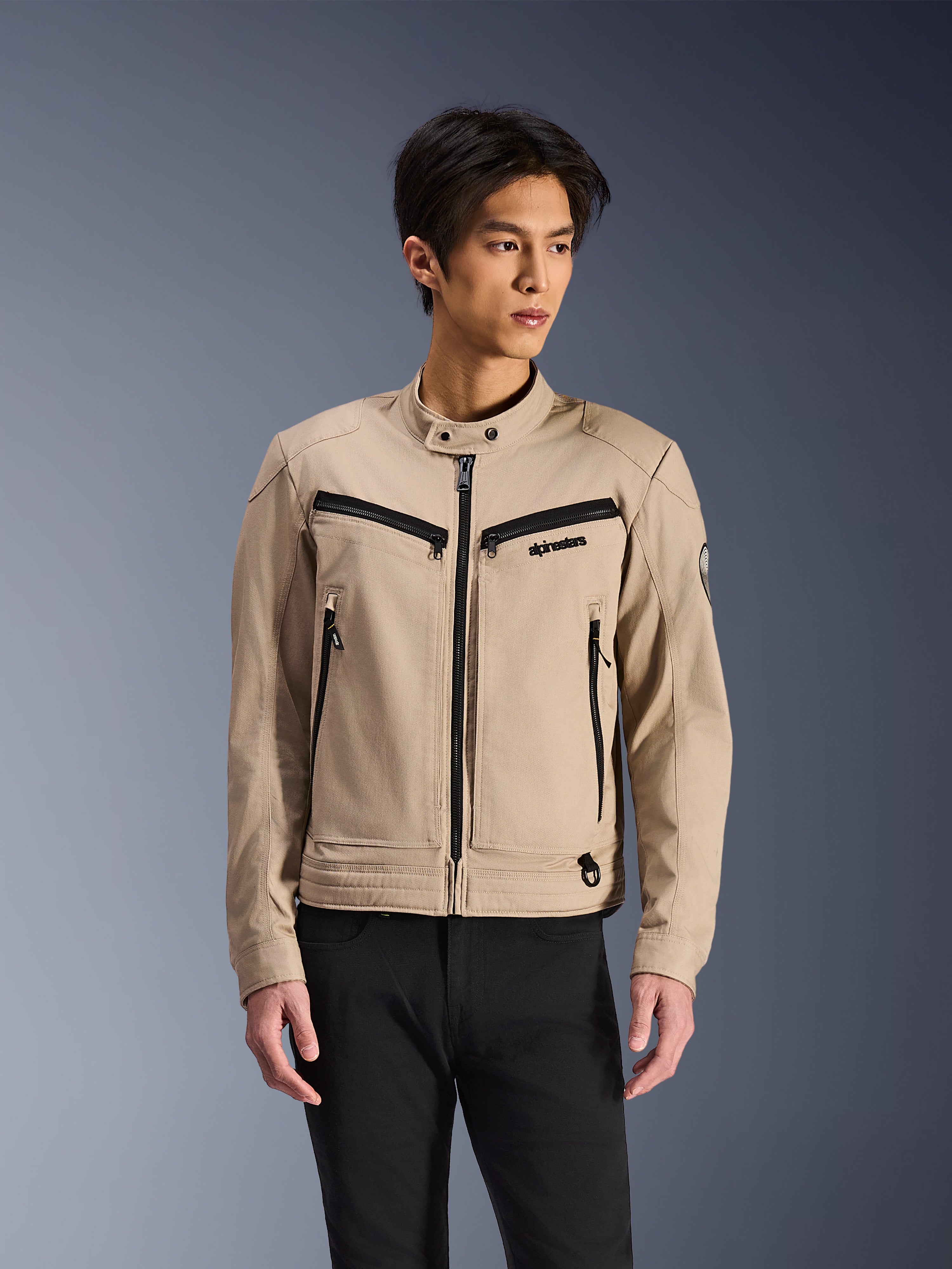 Alpinestars Bruiser Jacket, chaqueta de moto beige llevada por un modelo masculino, con detalles de cremalleras negras en el pecho y bolsillos laterales, cuello alto con botones a presión, prenda de conducción urbana inspirada en el estilo workwear, combinada con pantalones negros