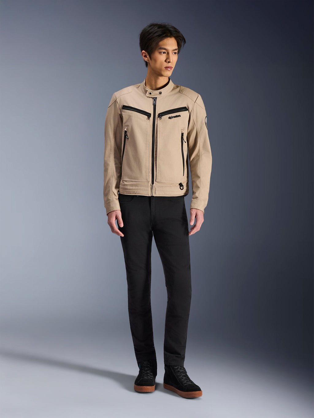 Chaqueta Alpinestars Bruiser, chaqueta urbana de motociclismo en color Aluminum, combinada con pantalones negros y zapatillas negras, con bolsillos negros con cremallera y cuello con botón a presión, diseño contemporáneo inspirado en la ropa de trabajo para conducción urbana