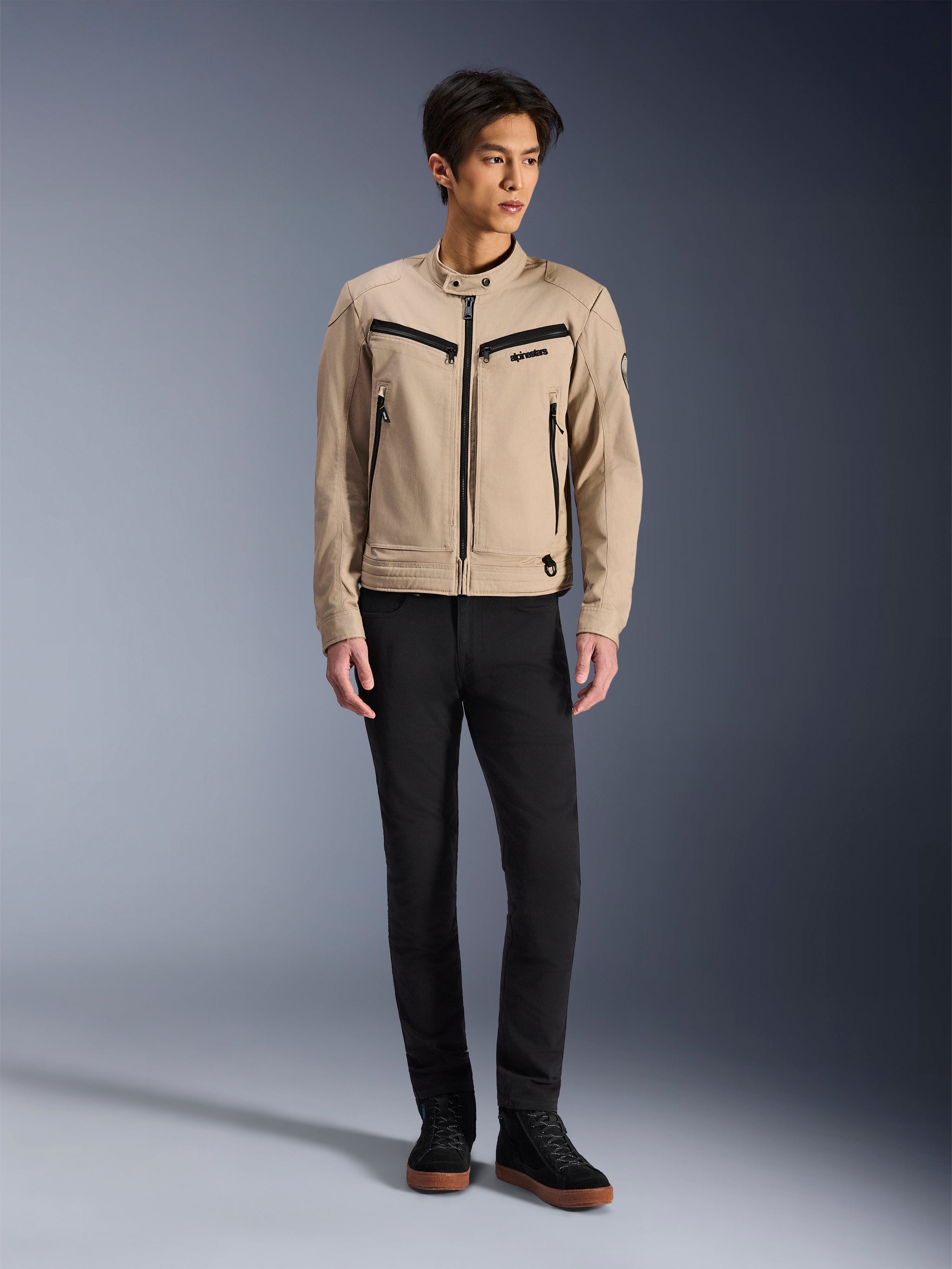 Chaqueta Alpinestars Bruiser, chaqueta urbana de motociclismo en color Aluminum, combinada con pantalones negros y zapatillas negras, con bolsillos negros con cremallera y cuello con botón a presión, diseño contemporáneo inspirado en la ropa de trabajo para conducción urbana