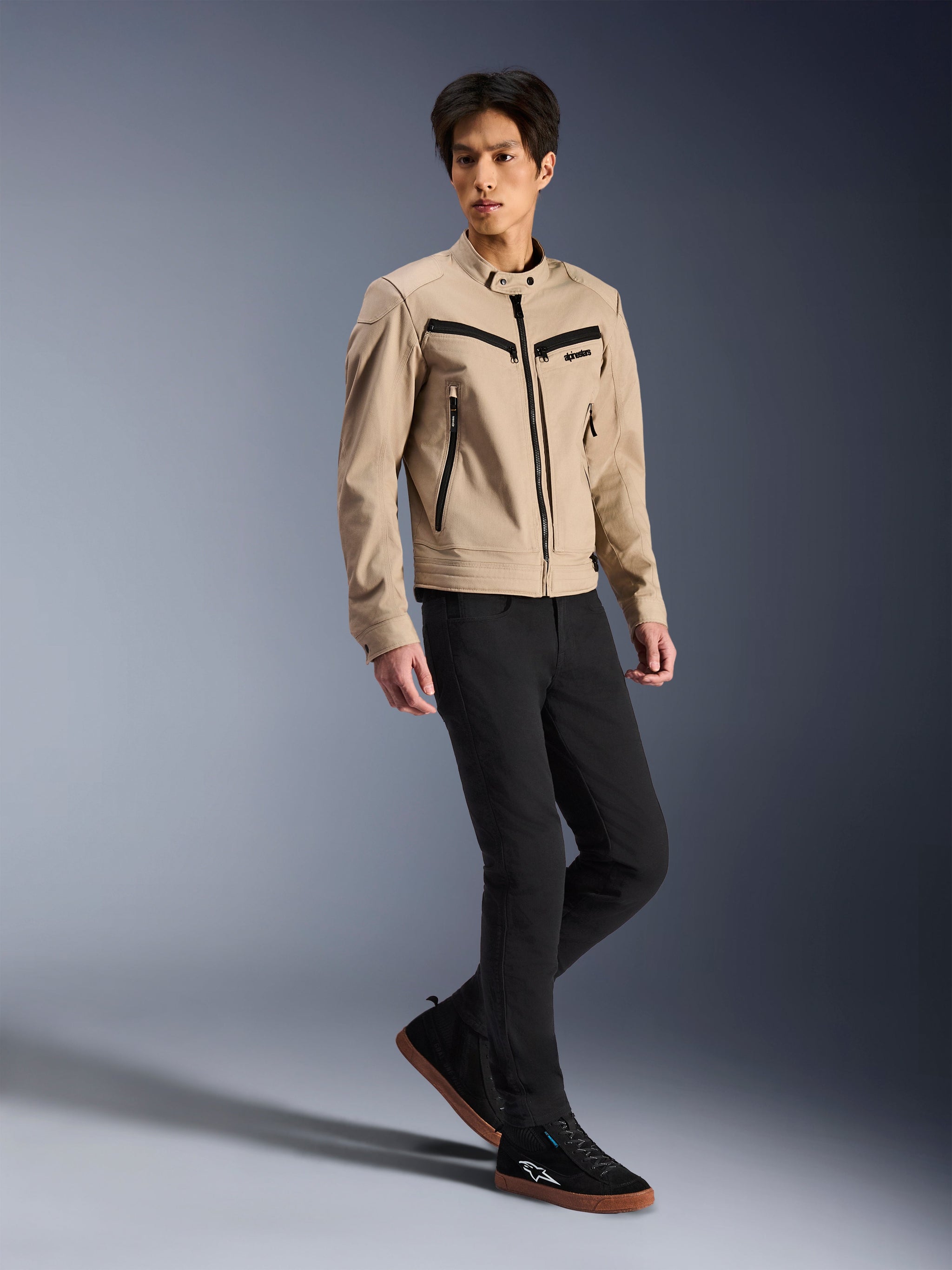 Chaqueta Alpinestars Bruiser, chaqueta urbana de motociclismo, color canela, lucida por un modelo masculino con pantalones negros ajustados y zapatillas negras, construcción textil inspirada en ropa de trabajo con detalles de cremalleras negras, estilo urbano contemporáneo.