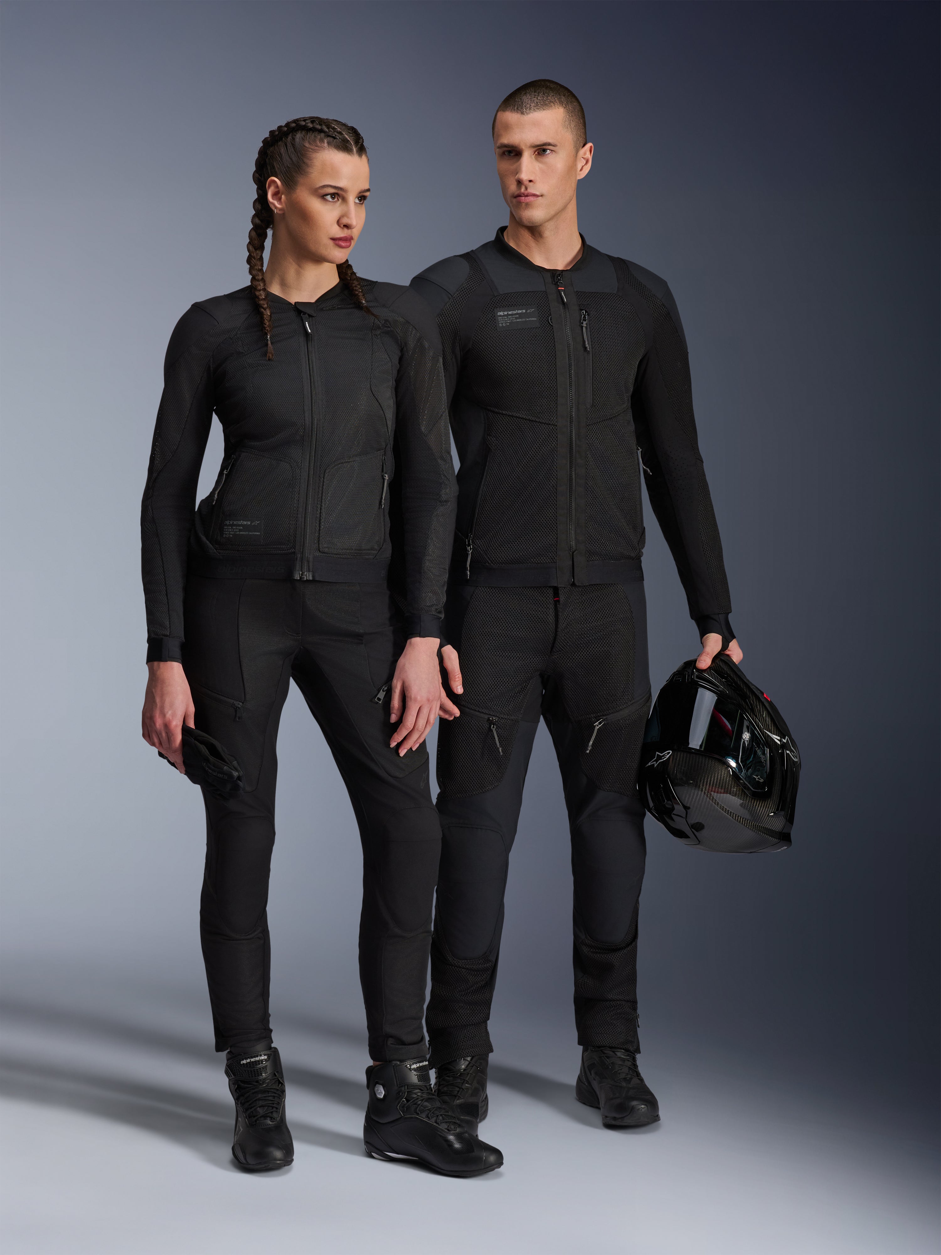 Chaqueta Alpinestars Stella Troop-Air, hombre y mujer con equipamiento técnico de motociclismo a juego en color negro, con construcción de malla para el flujo de aire, pantalones y botas negras, hombre sosteniendo un casco negro, ropa de conducción urbana